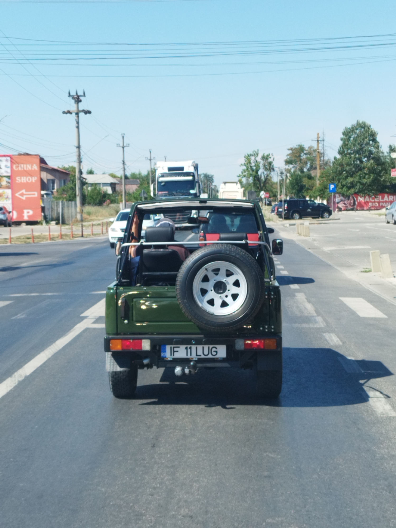 A green, open-top off-road vehicle is stopped at a crosswalk on a sunny day. The vehicle is in the foreground, facing away from the camera, and its license plate reads "IF 11 LUG". Behind the vehicle, a white truck is visible, along with other cars and buildings. A sign on the left side of the road says "CHINA SHOP". The scene appears to be in an urban or suburban area of Crevedia, Romania, with power lines and trees lining the street. The sky is clear and blue.