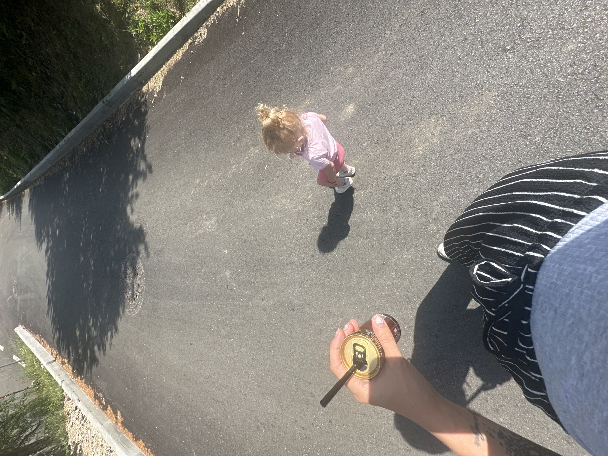 A low-angle, outdoor, eye-level shot captures a paved walkway bordered by a grassy slope and a curb. The walkway is mostly shaded by unseen trees, with dappled sunlight illuminating sections of the asphalt. In the distance, a young child with blonde hair pulled into a bun, wearing a pink shirt and shorts, walks away from the camera, casting a shadow. In the foreground, a person's hand holds a can with a straw, their arm and torso also visible, clad in a grey top and black-and-white striped pants. The scene appears to be set in a park or recreational area within the city of Tryavna, Bulgaria, possibly during a sunny day.