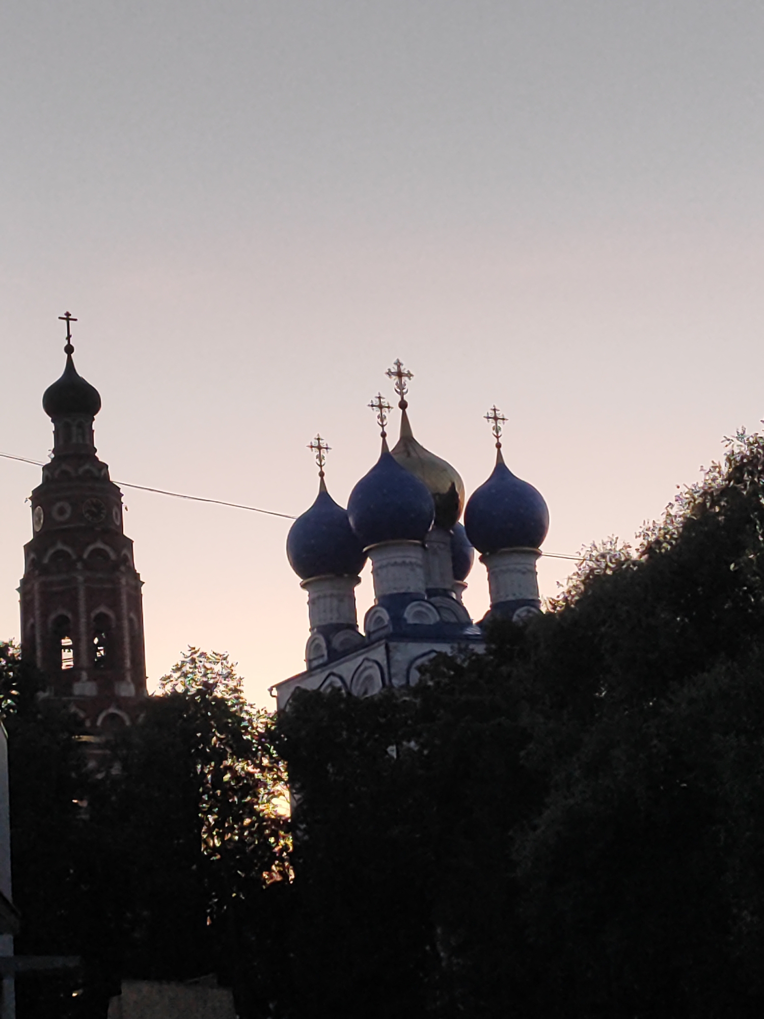 The image shows the bell tower and several domes of the Church of the Resurrection of Christ in Bronnitsy, Russia, against a pale sky at dusk. The bell tower, on the left, is tall and slender, constructed of red brick, with arched openings and a clock face visible. The domes of the church are a mix of blue and gold, topped with crosses. Dark green trees partially obscure the lower portions of the structures. A wire runs horizontally across the middle of the frame. The scene is largely devoid of people, focusing on the architecture and the atmospheric lighting.