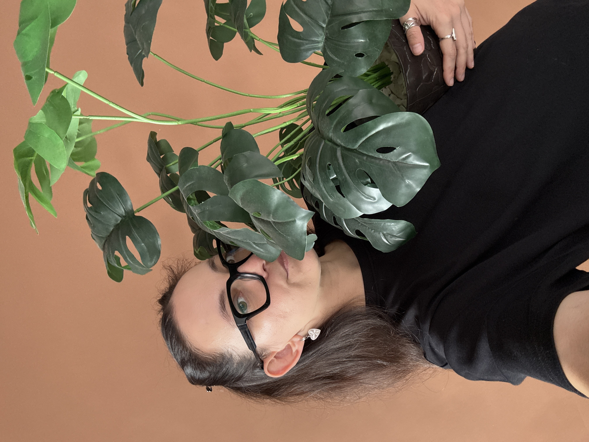 A medium shot shows a woman with dark hair pulled back in a ponytail, wearing black-rimmed glasses and a black t-shirt. She is holding a potted artificial Monstera plant in front of her. Her left hand is visible supporting the pot, adorned with two silver rings. Her right hand is not visible. She is looking towards the camera with a slight smile, her face partially obscured by the plant's large, green leaves. The background is a solid, light brown wall. The setting appears to be indoors, possibly a domestic interior, with no discernible urban context from Chaikovsky, Russia, or any other city. The lighting is soft and diffused, suggesting natural or studio lighting.