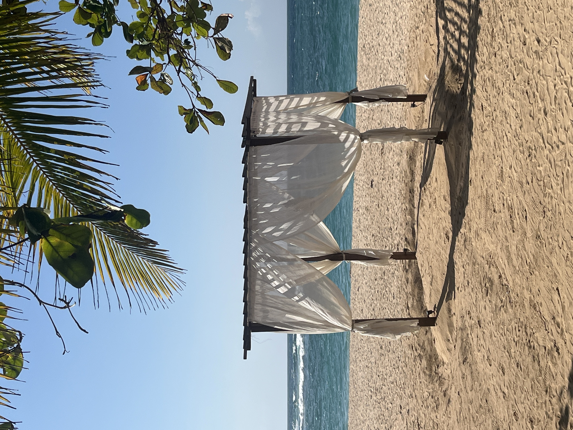 The image depicts a beach scene with a wedding arch in the foreground, adorned with white drapes. The arch is situated on a sandy beach facing a turquoise ocean with gentle waves. Lush green foliage from palm trees and other tropical plants frames the top of the image against a clear blue sky. The lighting suggests it is a sunny day, casting shadows from the arch onto the sand. This image is representative of many beautiful beach locations, including those found in Kuta Selatan, Indonesia, known for its picturesque coastlines and resorts. There are no people visible in the image.