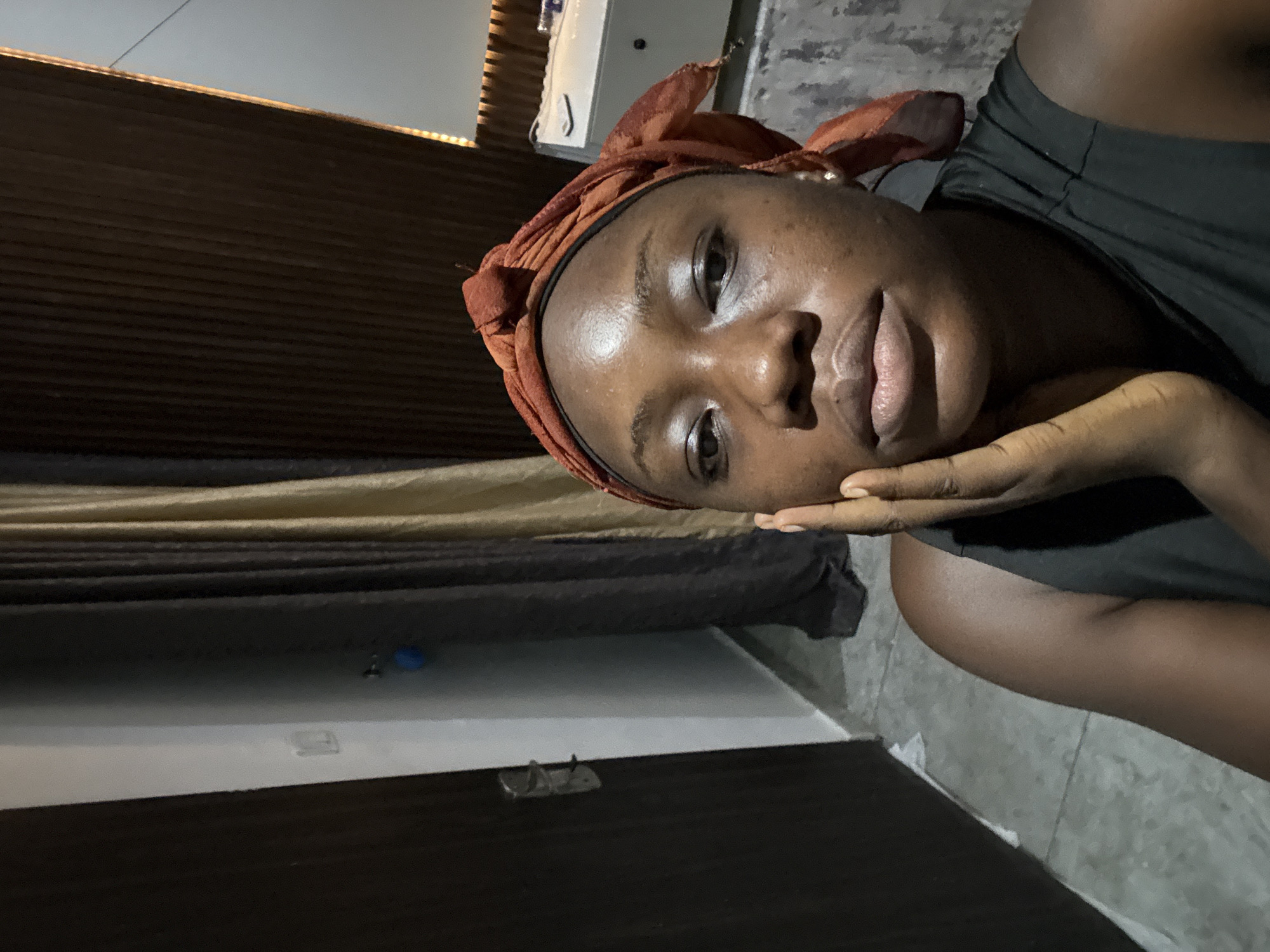 A young woman with dark skin and an auburn headscarf is the focal point of this indoor photograph. She rests her head in her hand, gazing at the camera with a soft expression. She is wearing a dark-colored sleeveless top. The location is Iddo Tudunwada, Nigeria. The background includes a wall with horizontal wood paneling in varying shades of brown and beige. A section of the floor is visible, along with a dark-colored door and light-colored tiling. Some objects are placed above the woman’s head. The lighting is subdued.