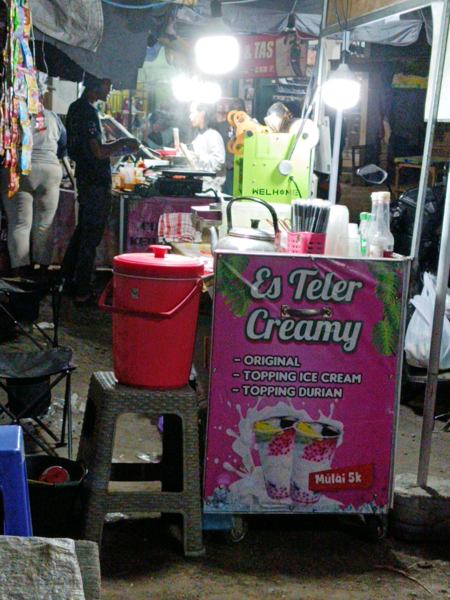 An outdoor food stall in Dompol, Indonesia, displays a vibrant pink banner advertising "Es Teler Creamy," with text indicating "Original," "Topping Ice Cream," and "Topping Durian." Two illustrated cups of the beverage are featured. A red cooler with a lid sits on a textured stool. Behind the cooler, a man wearing a dark shirt and a cap is preparing food at a counter. Another person, possibly female, stands at the counter facing the man. A green ice shaver machine with orange accents is visible, labeled "WELHOME." Various items, including bottles of condiments, a kettle, and utensils, are arranged on the stall. In the background, a large overhead tarp is supported by poles, and more stalls and lighting are suggested. A blue plastic stool is partially visible in the foreground to the left.