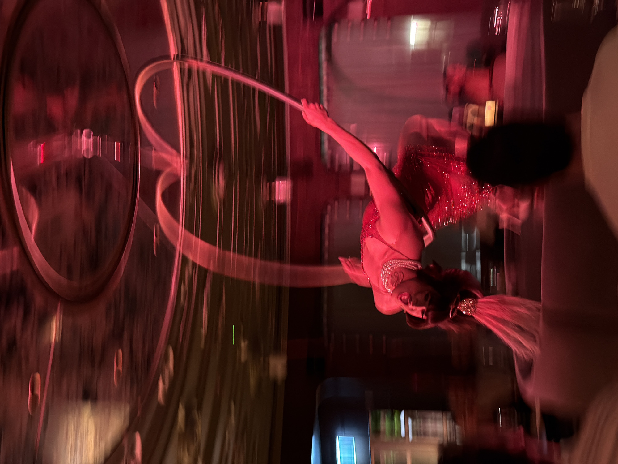 Captured in a dimly lit ambiance in Tel-Aviv, Israel, the image features a female performer, dressed in a red, sequined flapper-style dress, adorned with a pearl necklace, and a feathered headpiece, gracefully suspended from a hoop. She appears to be in the midst of an aerial performance, illuminated by reddish lighting. The background includes interior elements such as tall, vertical lines suggestive of architectural details, along with blurred figures indicating an audience or other individuals present in the venue. There are round elements visible on the left side of the image, possibly part of the performance equipment. The surroundings suggest an indoor performance or entertainment venue in Tel-Aviv.