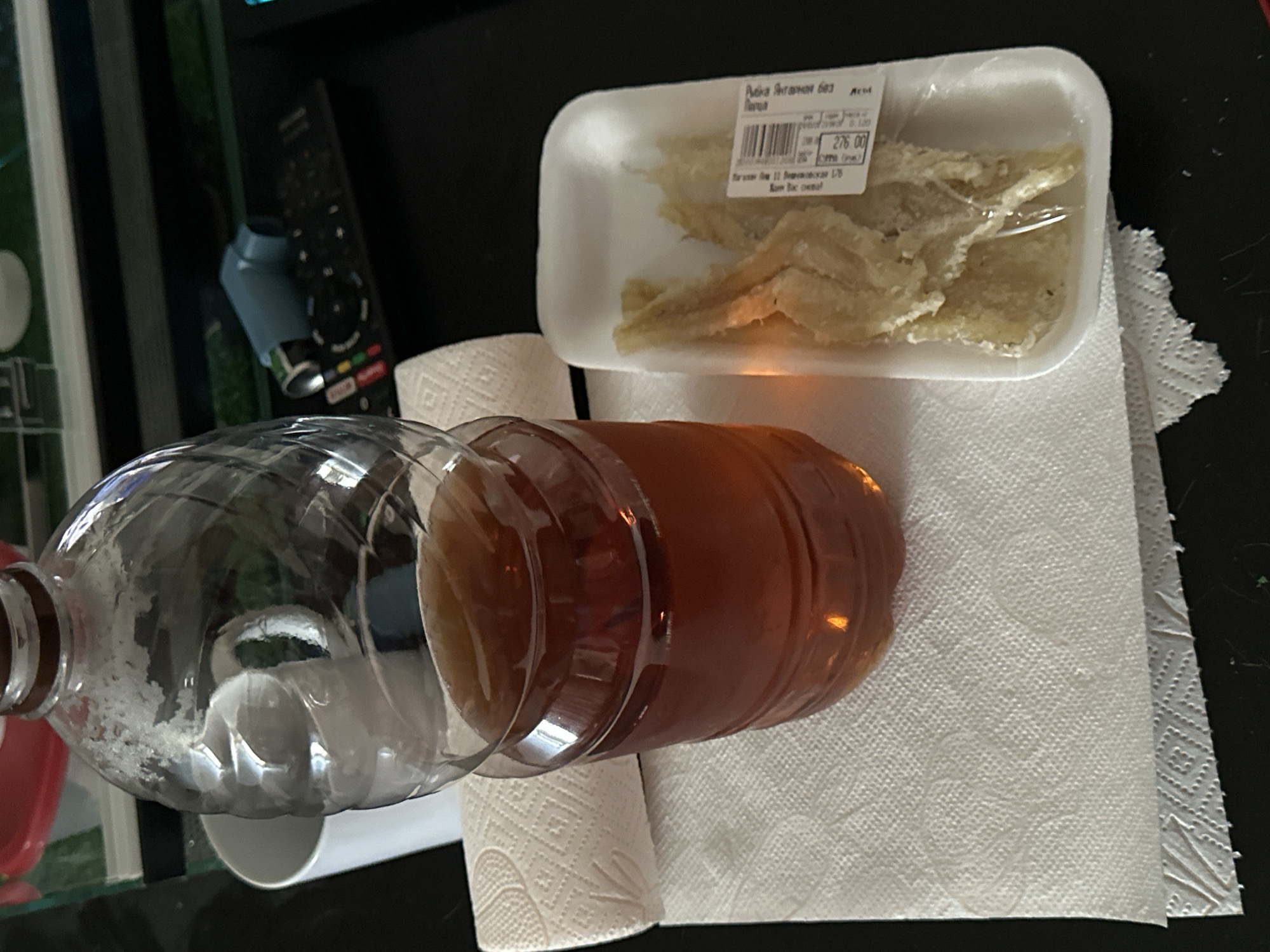 The image shows a still life of everyday items placed on a dark surface. A partially empty plastic bottle, filled with a brown liquid, rests on a roll of white paper towels. Next to it, on a sheet of paper towels, is a white styrofoam tray containing dried fish fillets wrapped in plastic. The price sticker on the fish reads "Рубка Янтарная 603 Кета" with a price of 276.00. In the background, there is a television remote control and a partially visible object resembling an inhaler. Given the product name "Рубка Янтарная" (Rubka Yantarnaya), which means "Amber Cut," and the fish being "кета" (keta), which is chum salmon, it is plausible that the image was taken in a location where Russian is spoken, potentially in Moscow, Russia.