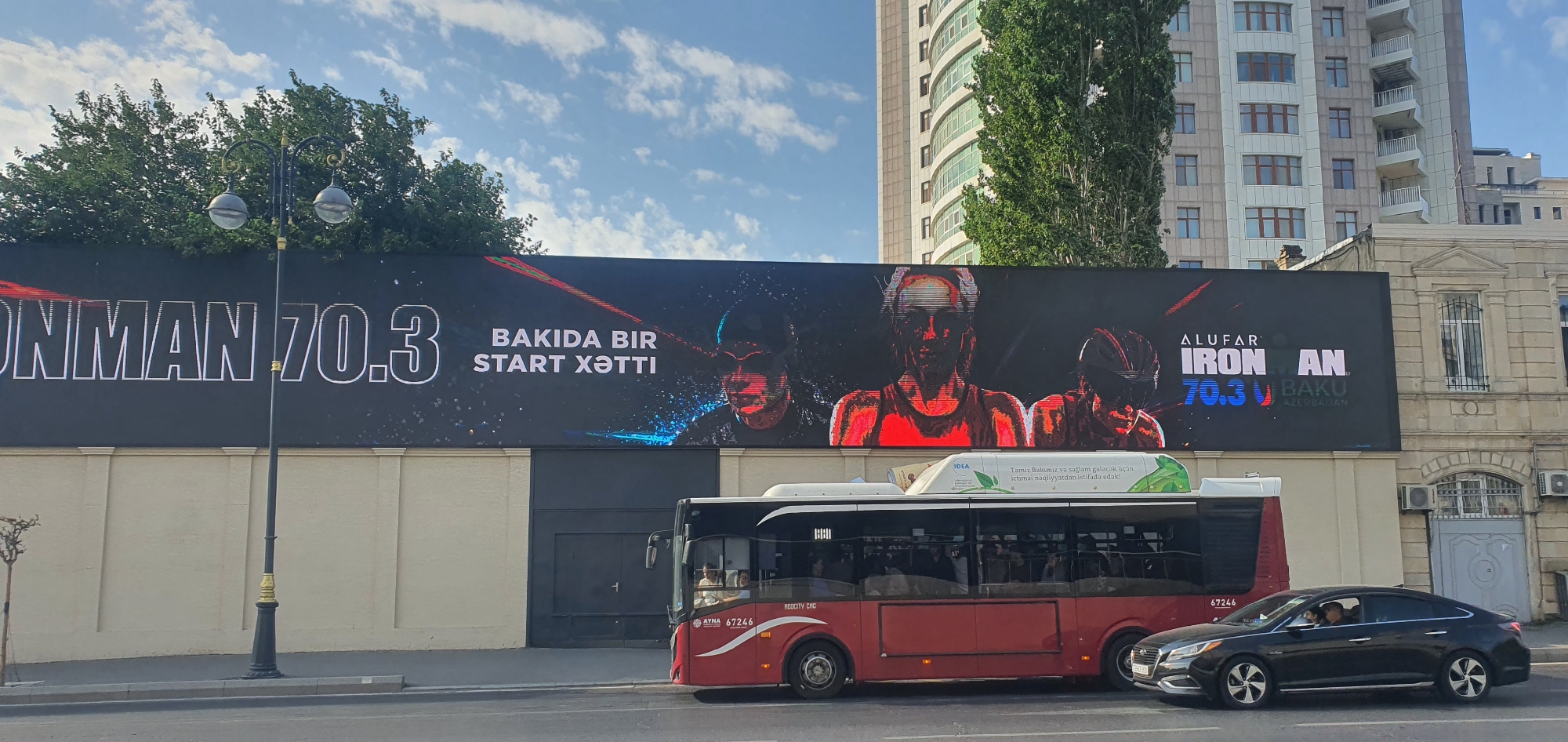 The image shows a large billboard displaying an advertisement for "ALUFAR IRONMAN 70.3 BAKU AZERBAIJAN." The billboard features stylized portraits of athletes, likely triathletes, and text in both English and Azerbaijani. Text on the billboard reads "ONMAN 70.3" and "BAKIDA BIR START XƏTTİ," which translates to "One Start Line in Baku."
In front of the billboard, a red bus with the number "67246" on its side is parked on the street. To the right of the bus, a black sedan is visible, also on the street. The bus has advertisements on its side, including one with the word "IDEA" and text in Azerbaijani related to health and transportation.
The environment appears to be an urban setting in Baku, Azerbaijan. Behind the billboard, a modern, tall building with a curved facade is visible, along with trees and a portion of an older, ornate building. A street lamp with two spherical lights is positioned in front of the billboard. The sky is partly cloudy.