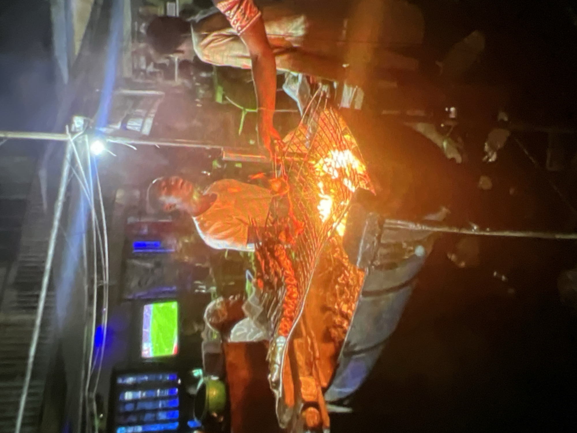 In Lagos, Nigeria, at night, people are gathered around a large barbecue grill that is actively cooking meat. The grill appears to be made from a repurposed barrel. Several skewers of meat are being cooked over an open flame, with visible embers and smoke. A young man in an orange shirt stands near the grill, and another person, partially visible, is tending to the meat. In the background, a television screen displays a soccer game. The environment suggests an outdoor setting, possibly a street food stall or a casual gathering place, illuminated by artificial lights.