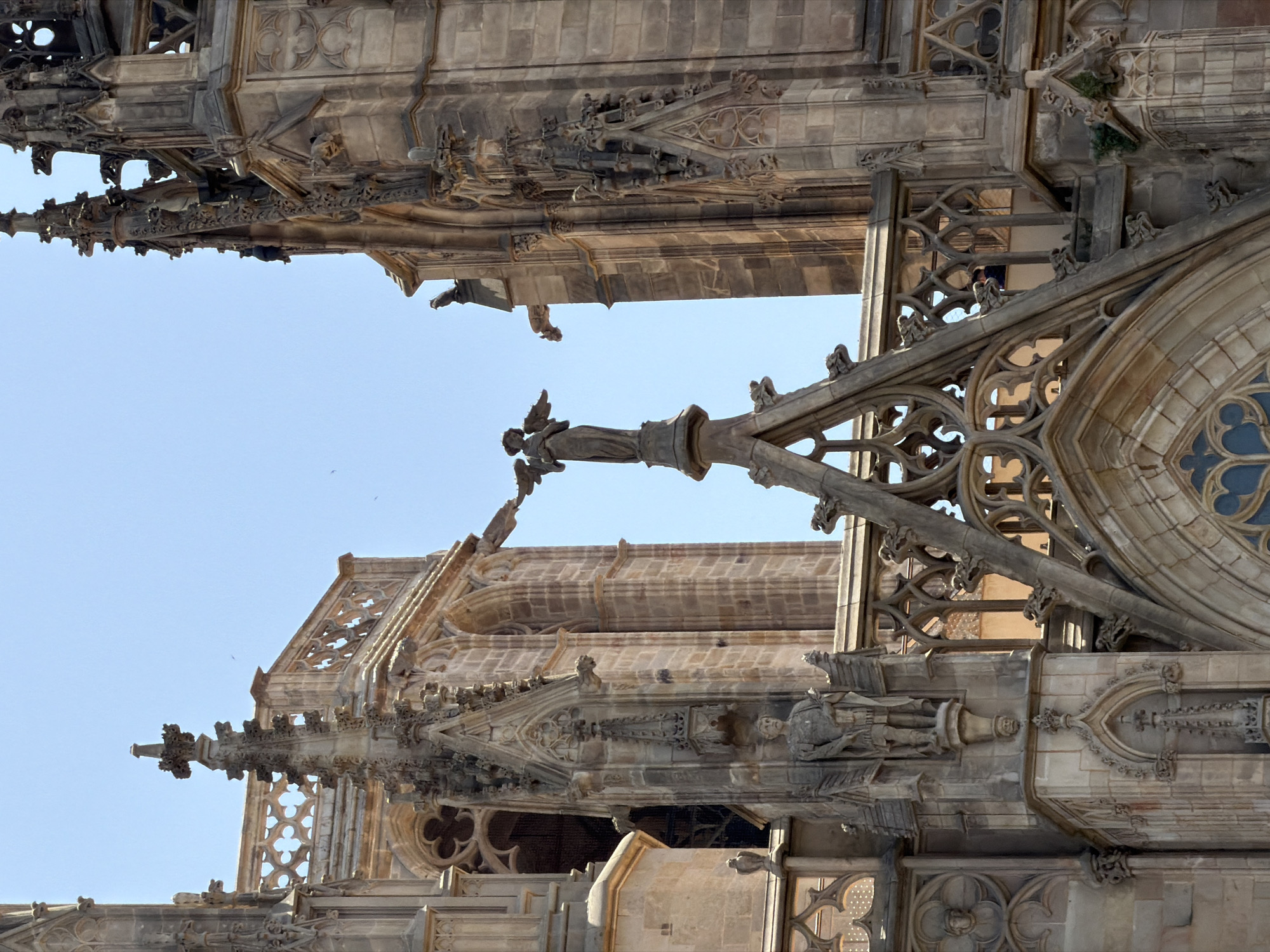 The image shows a close-up view of the exterior architecture of the Barcelona Cathedral, also known as the Cathedral of the Holy Cross and Saint Eulalia, located in the Gothic Quarter of Barcelona, Spain. The scene is dominated by ornate stone carvings, Gothic spires, and arched windows, characteristic of the cathedral's medieval design.
In the center of the frame, atop a pointed spire, stands a statue of an angel with outstretched wings. The angel is rendered in a dark stone and appears to be the focal point of the composition. To the left of the angel, on a protruding section of the cathedral, is a stone statue of a male figure, possibly a saint or historical figure, dressed in a tunic and holding an object in his right hand. This statue is positioned within a niche supported by a decorative bracket, below which is another smaller carved head.
The cathedral's façade is constructed from weathered, light-colored stone blocks. Intricate stonework adorns the spires and edges, including decorative tracery, gargoyles, and smaller sculpted figures. The sky behind the cathedral is a clear, pale blue.
The architectural elements suggest a grand and ancient religious structure, with a focus on verticality and detailed craftsmanship. The lighting indicates it is daytime, with bright sunlight casting shadows on the intricate carvings. The perspective is from below, looking up towards the upper portions of the cathedral, emphasizing its height and imposing presence.