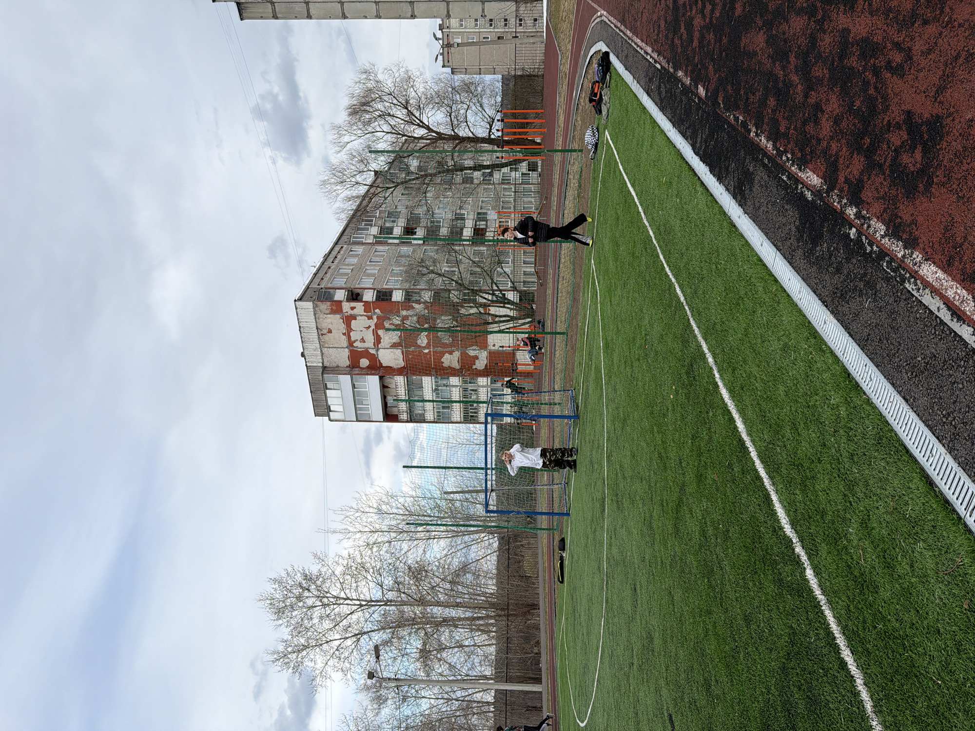 The image displays an outdoor sports complex featuring a green artificial turf field and a reddish-brown running track, set against an urban backdrop. The scene is captured on an overcast day, likely during a cooler season, as evidenced by the bare trees scattered around the area.
In the foreground, the well-maintained artificial turf field is marked with white lines, typical for soccer or football. A metallic drainage grate separates the turf from the adjacent running track. Sports equipment, including bags and jackets, are casually placed on the track.
The midground features two visible individuals on the field. One person, dressed in a white top and camouflage-patterned pants, stands inside a blue-framed goal with a net. Another individual, wearing a dark jacket and dark pants with distinct yellow-soled shoes, is seen walking or standing on the turf. Bare trees provide a natural element, some appearing behind a chain-link fence.
In the background, several multi-story residential buildings dominate the skyline. One building prominently features worn facades with significant red and white peeling paint, suggesting age. A taller, more uniform building is visible further back. Overhead, power lines traverse the cloudy, light grey sky.
The activity appears to be recreational sports, possibly a casual game or practice. The general setting is consistent with a public or school sports facility in a residential area. The architectural style of the buildings and the overall environment are indicative of the provided location context of Berezniki, Russia. No visible text is legible in the image.