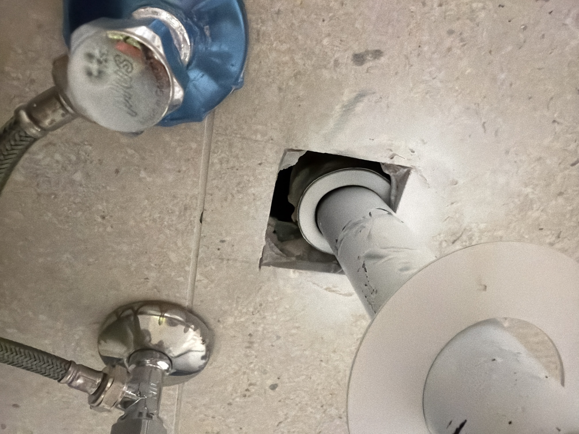 The image shows plumbing under a sink, likely in a bathroom or kitchen. Two water supply lines are visible, one connected to a chrome shut-off valve and another to a larger, blue-covered valve. A section of exposed pipe extends from a hole in the tiled wall, with a white flange or escutcheon plate visible. The wall appears to be made of light-colored, textured tiles. There are no people or animals present. The scene is indoors, and the lighting suggests it is likely artificial. The overall impression is one of a functional plumbing setup, possibly in need of repair or maintenance. The provided location of Jerusalem, Israel, does not provide any specific visual cues that can be identified from the image itself.