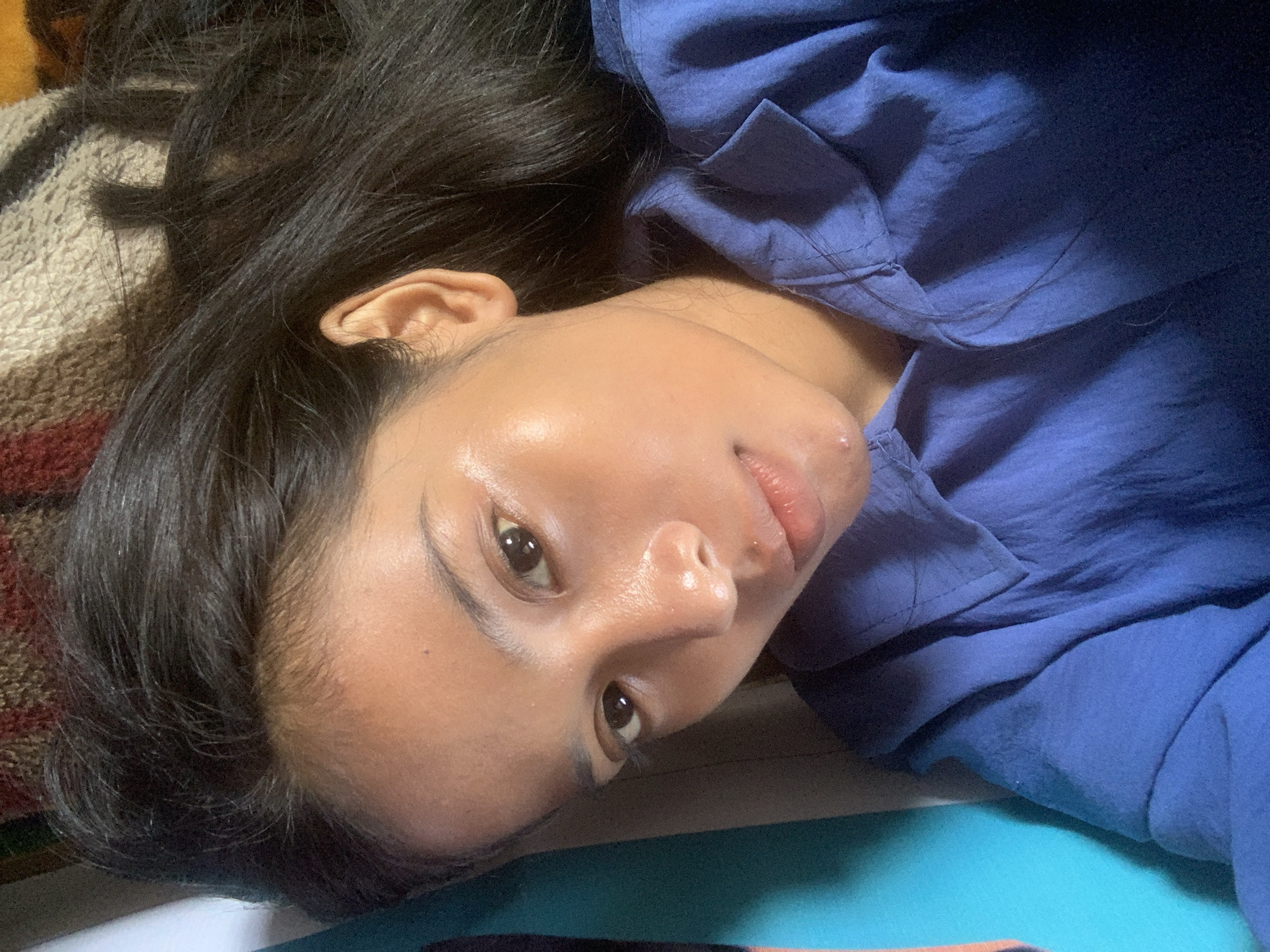 A close-up, high-angle portrait features a young individual, likely female, lying down and looking directly into the camera. The person's long, dark, straight hair is spread out around their head. They are wearing a blue, collared shirt made of a lightweight, possibly linen-like fabric, which shows some natural folds and a faint discoloration mark on the lower right.
The individual's skin appears smooth with a slight sheen, particularly on the forehead and cheeks, suggesting natural oils or a subtle glow. Their eyes are dark brown, and their lips are full with a natural, slightly rosy hue, featuring a small blemish on the upper lip. The expression is neutral and calm.
The setting appears to be indoors. To the left and behind the person's head, a textured, patterned fabric in shades of brown, beige, and red is visible, possibly a blanket or throw. To the right and below, a solid turquoise-blue surface, likely a bedsheet or another piece of fabric, provides a contrasting background. The lighting is soft and even, without strong shadows, indicating an indoor environment. There is no visible text or specific activity beyond the individual resting and posing for the photograph. The provided context places this scene in Sungai Meriam, Indonesia.