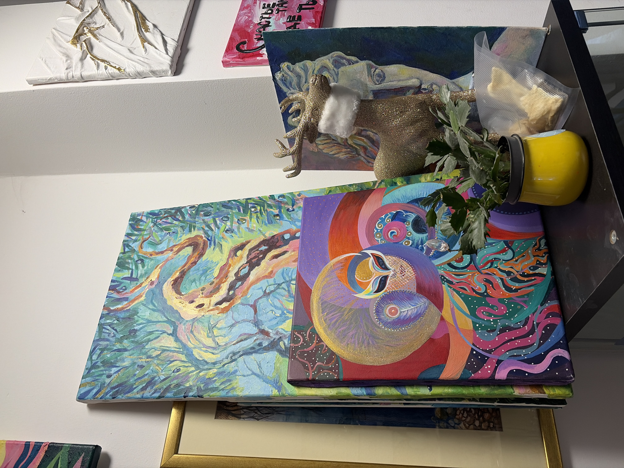 The image presents a cluttered shelf space. On the top shelf, there are several paintings, including a white textured painting with gold accents, a small painting with pink text, and a landscape painting with a reclining figure. A gold glitter reindeer figurine with a white cotton scarf and a vacuum-sealed bag containing white objects are also present. On the lower shelf, there are several paintings stacked on top of each other. A painting with a swirling, colorful design and another with a more naturalistic landscape are visible. A yellow planter containing a plant is leaning against the shelf's edge. A painting is also framed in gold on the bottom shelf. There is nothing in the image to indicate the location is Budva, Montenegro.