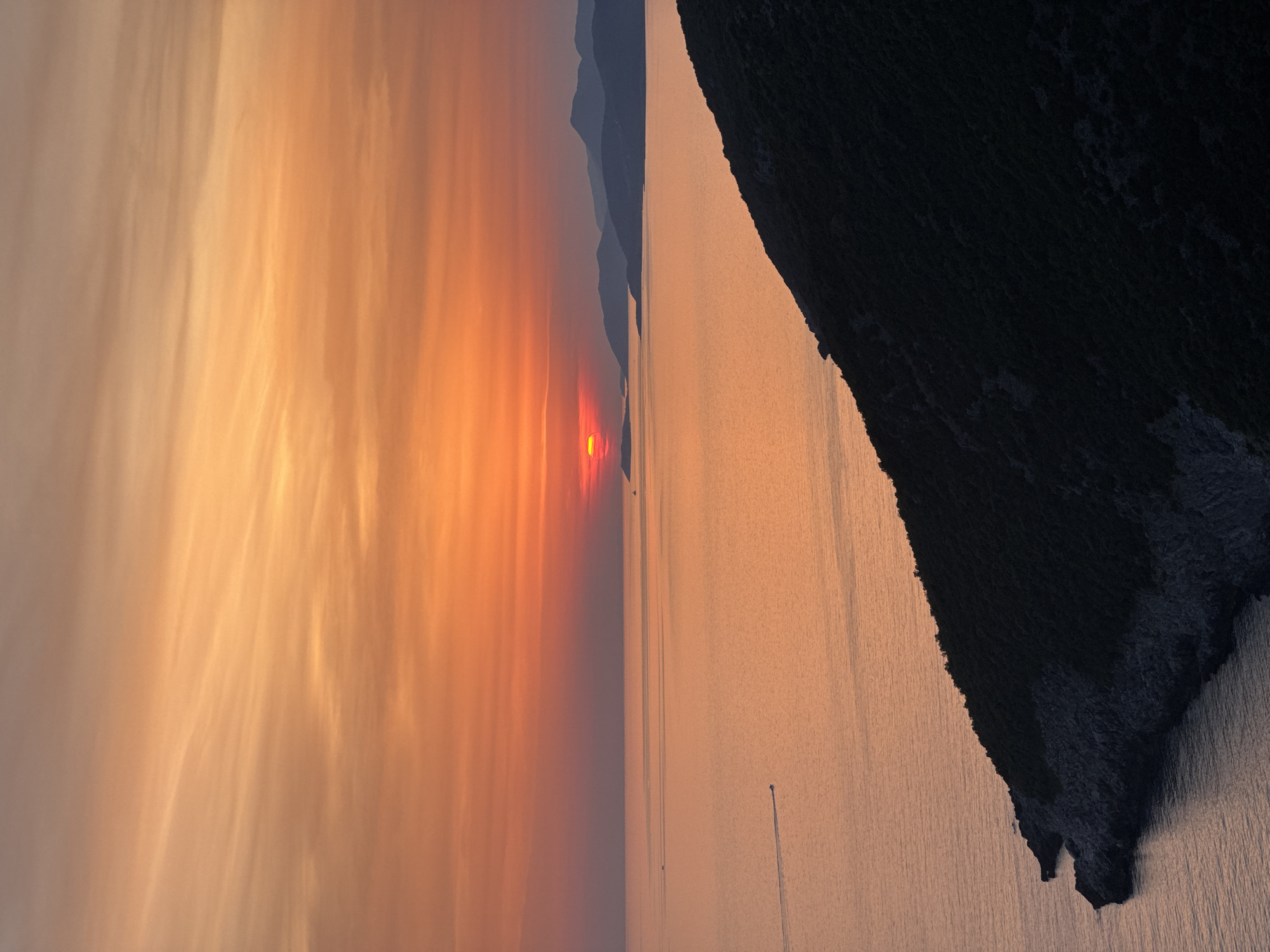 A vertical, high-angle shot captures a tranquil sunset over the Adriatic Sea, with the coast of Zagora, Montenegro, silhouetted in the foreground and distance. The sky is a gradient of warm oranges and yellows, with wispy clouds streaking horizontally. The sun, a bright orange disc, is partially submerged at the horizon, casting a golden glow across the calm water. In the middle distance, several islands or peninsulas are visible as dark, hazy shapes against the sunset. A small, dark boat with a visible wake is moving from left to right across the frame on the water. The immediate foreground is dominated by a steep, dark hillside covered in dense vegetation, with rocky outcrops visible near the water's edge. The overall impression is one of serene natural beauty.