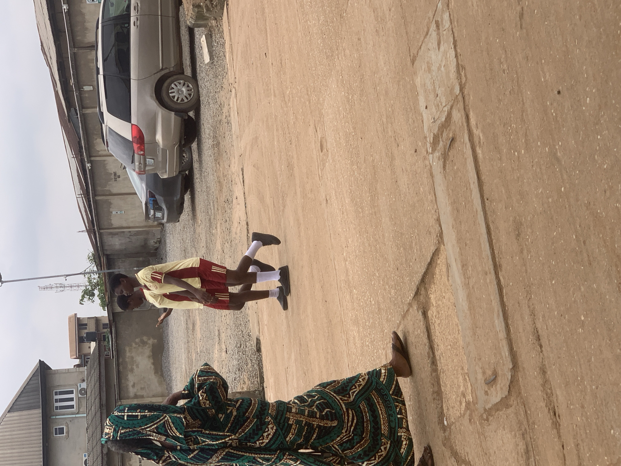 In Shagamu, Nigeria, on a cloudy day, a woman in a green and gold patterned kaftan walks on a dirt path. Behind her, two young people, dressed in yellow and red sports uniforms, walk in the same direction. They appear to be talking. In the background, a tan minivan and a dark car are parked on a gravel lot next to a long, gray concrete building with a corrugated metal roof. Buildings and a telecommunications tower are visible further in the distance.