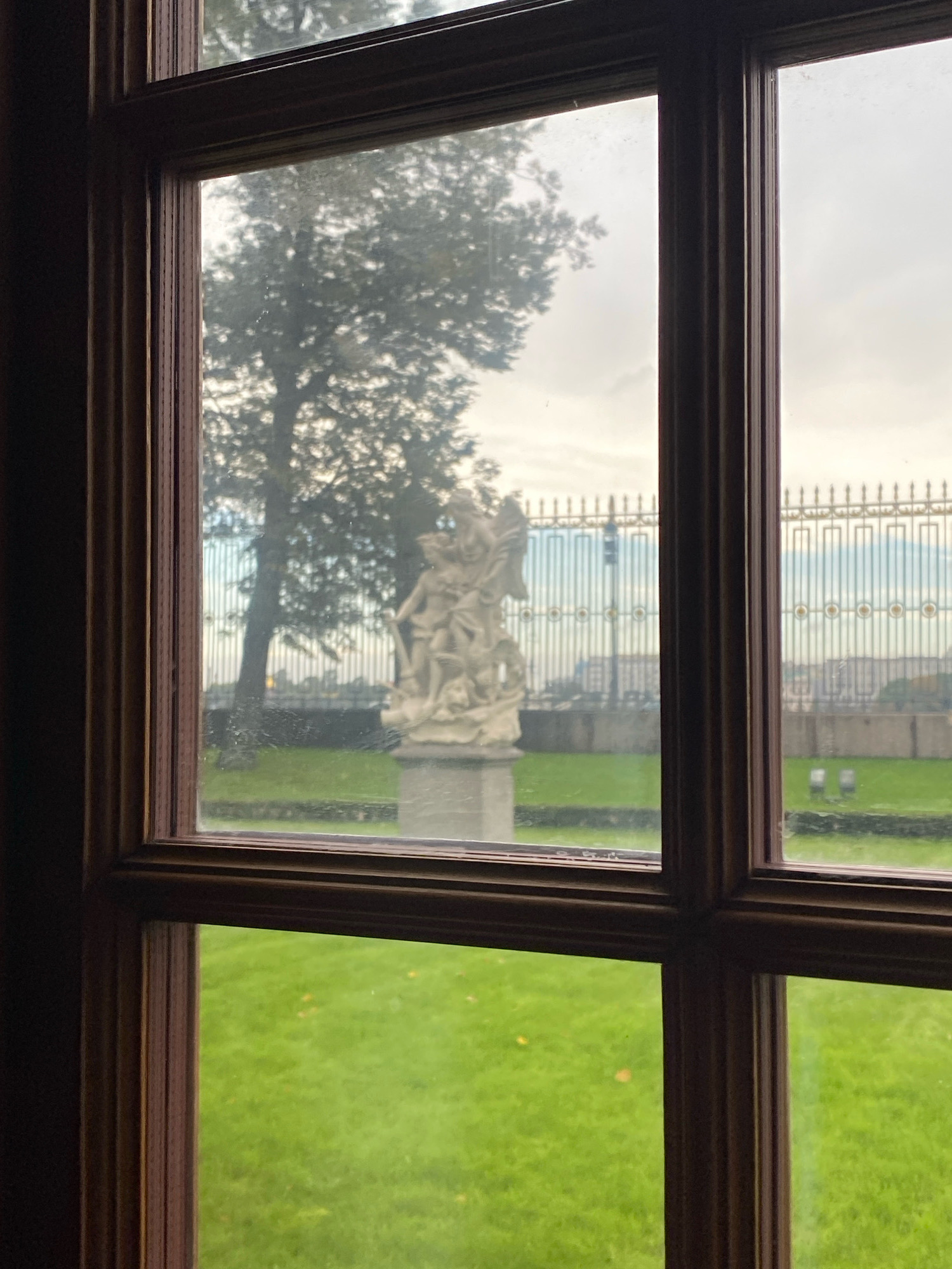 The image shows a view through a multi-pane window with dark wooden frames. In the foreground, the interior of a room is visible to the left of the window. Through the window, a lawn with scattered yellow leaves is seen in the lower half. In the middle ground, a stone sculpture of two human figures is positioned on the lawn. Behind the sculpture, a metal fence with ornate golden detailing divides the lawn from a background scene. Beyond the fence, a cityscape with buildings is visible, and beyond that, a body of water, likely a river, can be discerned. The sky is overcast. This scene is consistent with a view from the Winter Palace, looking out over Palace Square and the Admiralty Embankment towards the Neva River in Saint Petersburg, Russia.