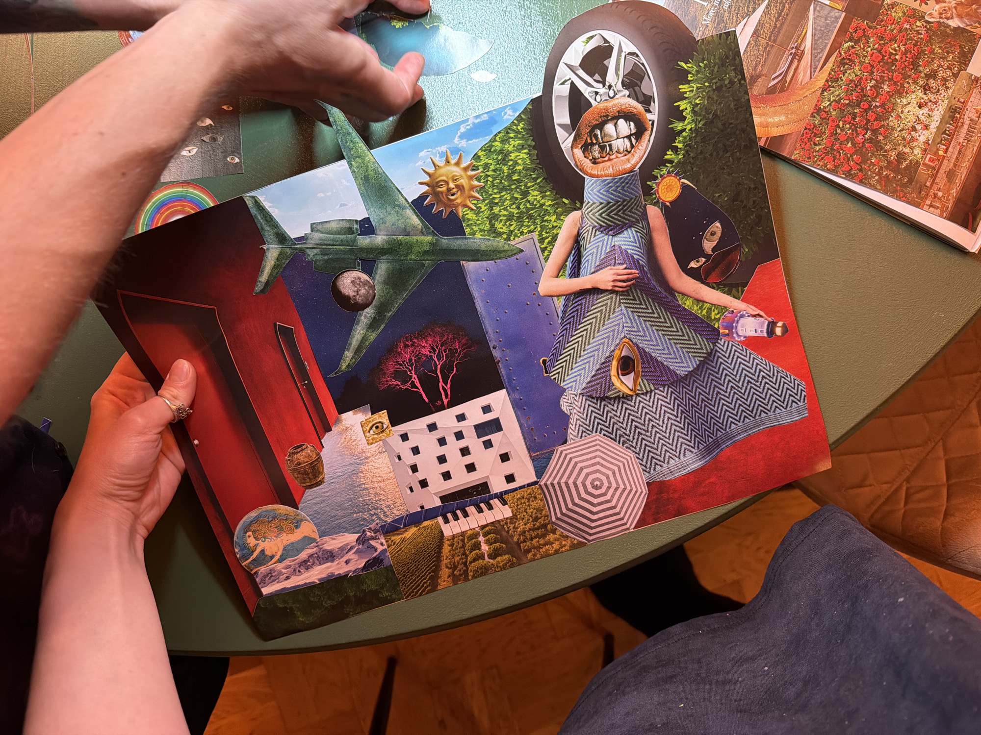 A collage is being created on a green round table. The collage contains a mix of images, including an airplane, a car tire with a mouth and teeth, a woman in a dress, buildings, and nature scenes. Two hands are visible, presumably belonging to the person creating the collage. Other cut-out images are on the table beside the collage. The location is indoors on a chair.