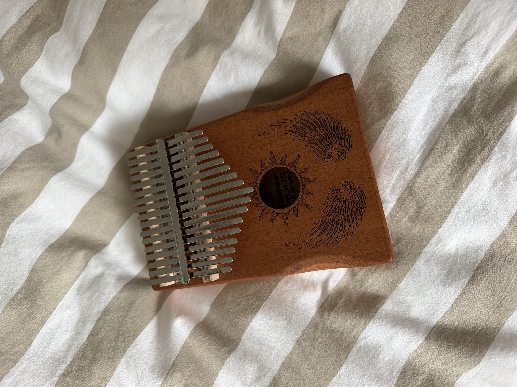 A Kalimba, also known as a thumb piano, is shown in the image, resting on a striped fabric surface. The Kalimba is made of wood and has metal tines attached to it. The wood has an engraved design of a sun with rays and a pair of wings. The striped fabric is composed of alternating beige and white stripes, which appear to be part of bedding or a tablecloth. The Kalimba is positioned diagonally across the frame. No people are visible in the image. The scene is indoors. The location of this image is not in Herne, Germany.