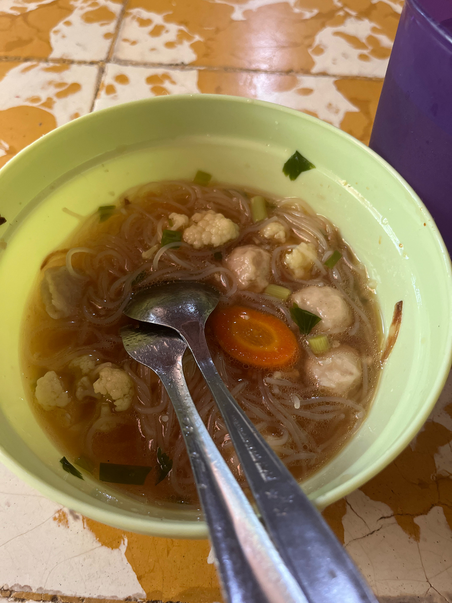 A close-up overhead shot shows a light green bowl filled with a noodle soup dish, positioned on a tiled surface. The soup contains thin, white noodles, several round meatballs, pieces of cauliflower, sliced carrots, and chopped green onions. Two metal spoons are resting inside the bowl, partially submerged in the broth. To the right of the bowl, a portion of a purple plastic cup is visible. The tiled surface beneath the bowl is yellow and white, with visible grout lines and some apparent wear. The overall setting appears to be a casual dining environment, possibly a local eatery in the city of Lambaro Kueh, Indonesia.