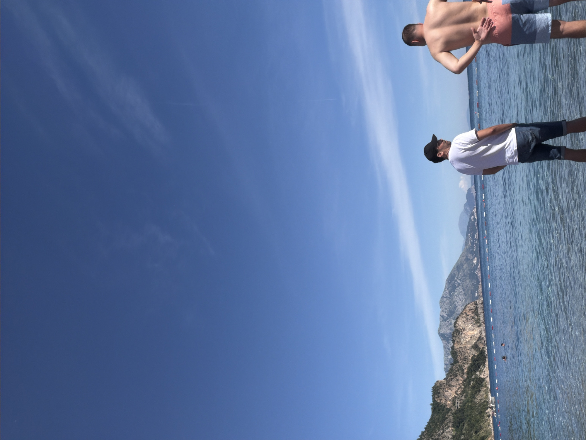 The image shows a coastal scene, likely in or near Prijevor, Montenegro, with a clear blue sky dominating the left side of the frame. Two men are visible along the shoreline. One man, wearing a white t-shirt, dark shorts, and a baseball cap, stands with his back to the camera. Another man, shirtless and wearing patterned swim trunks, is slightly further along the shore. The water is calm and appears to be shallow. A row of floats demarcates a swimming area. Rocky, forested mountains are visible in the background.