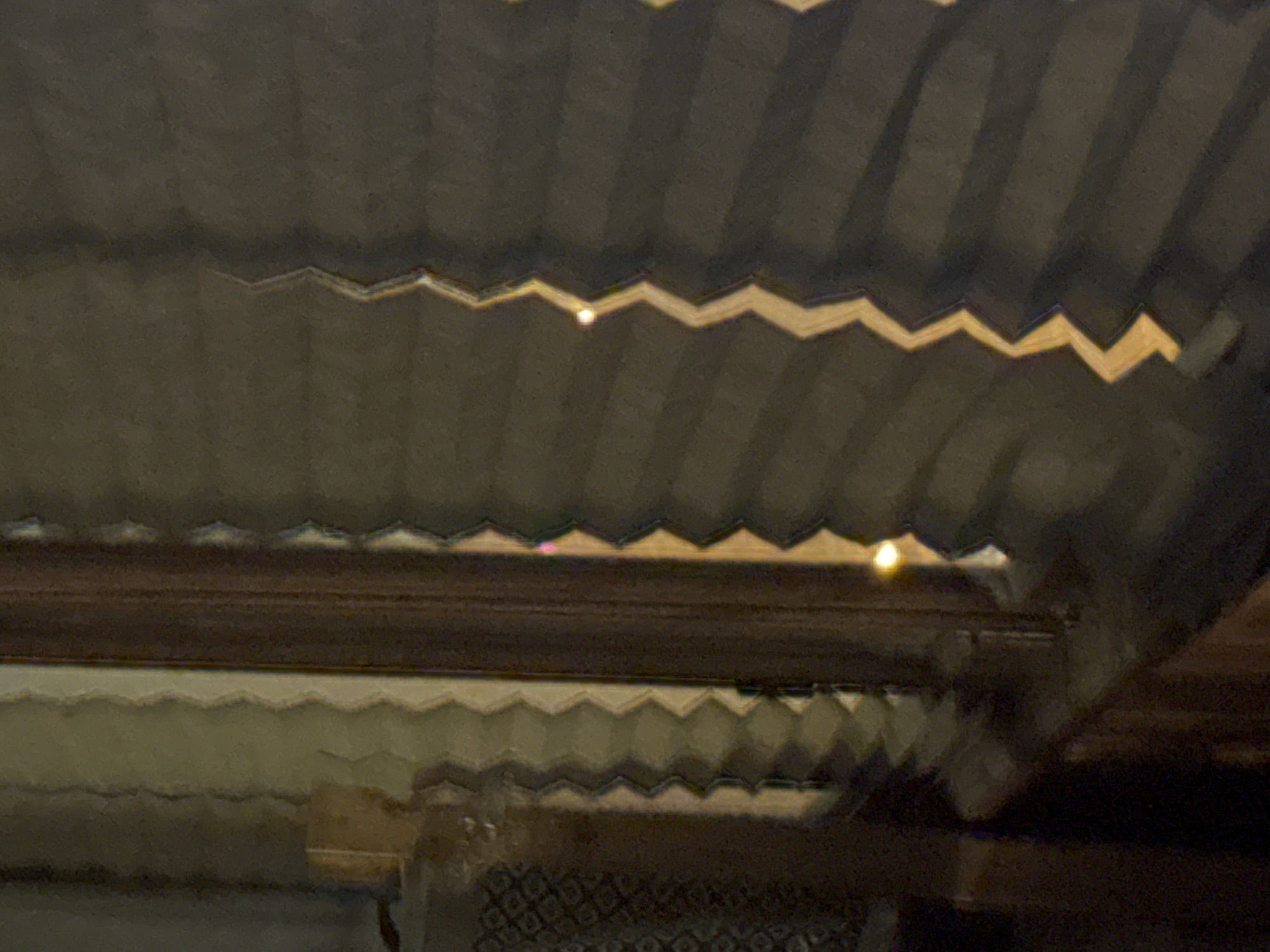 A close-up, somewhat blurry image reveals a building interior. The ceiling is structured with what appears to be rows of saw-tooth like architectural features, possibly skylights or ventilation shafts. These features are illuminated, suggesting sunlight is filtering through. Below the ceiling is what appears to be a dark-stained wooden beam, and underneath that, another row of saw-tooth architectural details. Below these details, more architectural features are visible. The image lacks clarity, but suggests the interior of a building in Krasnoyarsk, Russia.