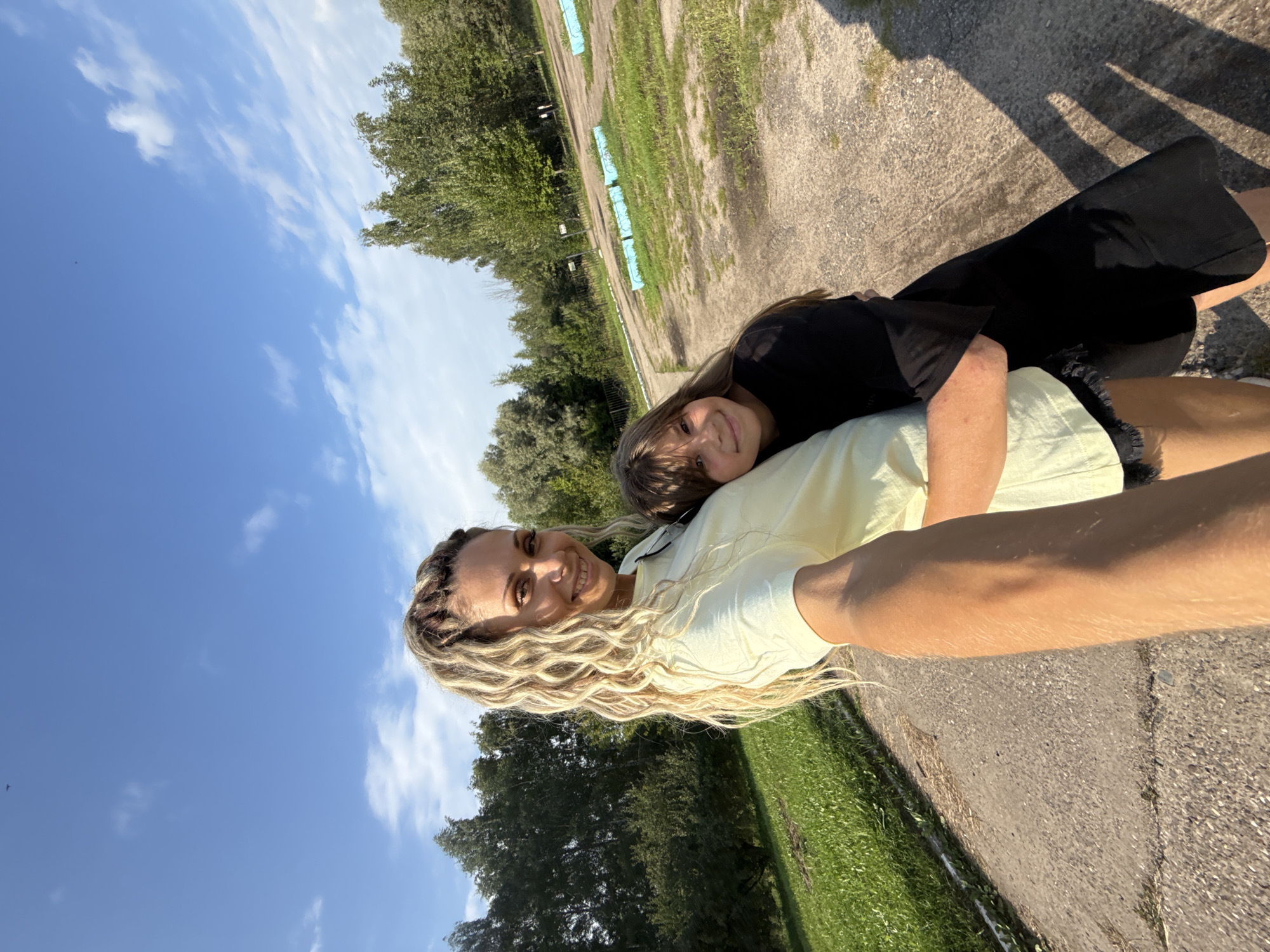 In Rusanovo, Russia, on a sunny day, a woman with blonde braided hair and a light yellow shirt embraces a young girl wearing a black t-shirt. They are standing on a paved path surrounded by green grass and trees. The sky is blue with scattered white clouds. The scene appears to be an outdoor recreational area, possibly a park or a sports field, with a fence visible in the background and some blue-covered benches in the distance. The woman is holding a phone, indicating they are taking a selfie.