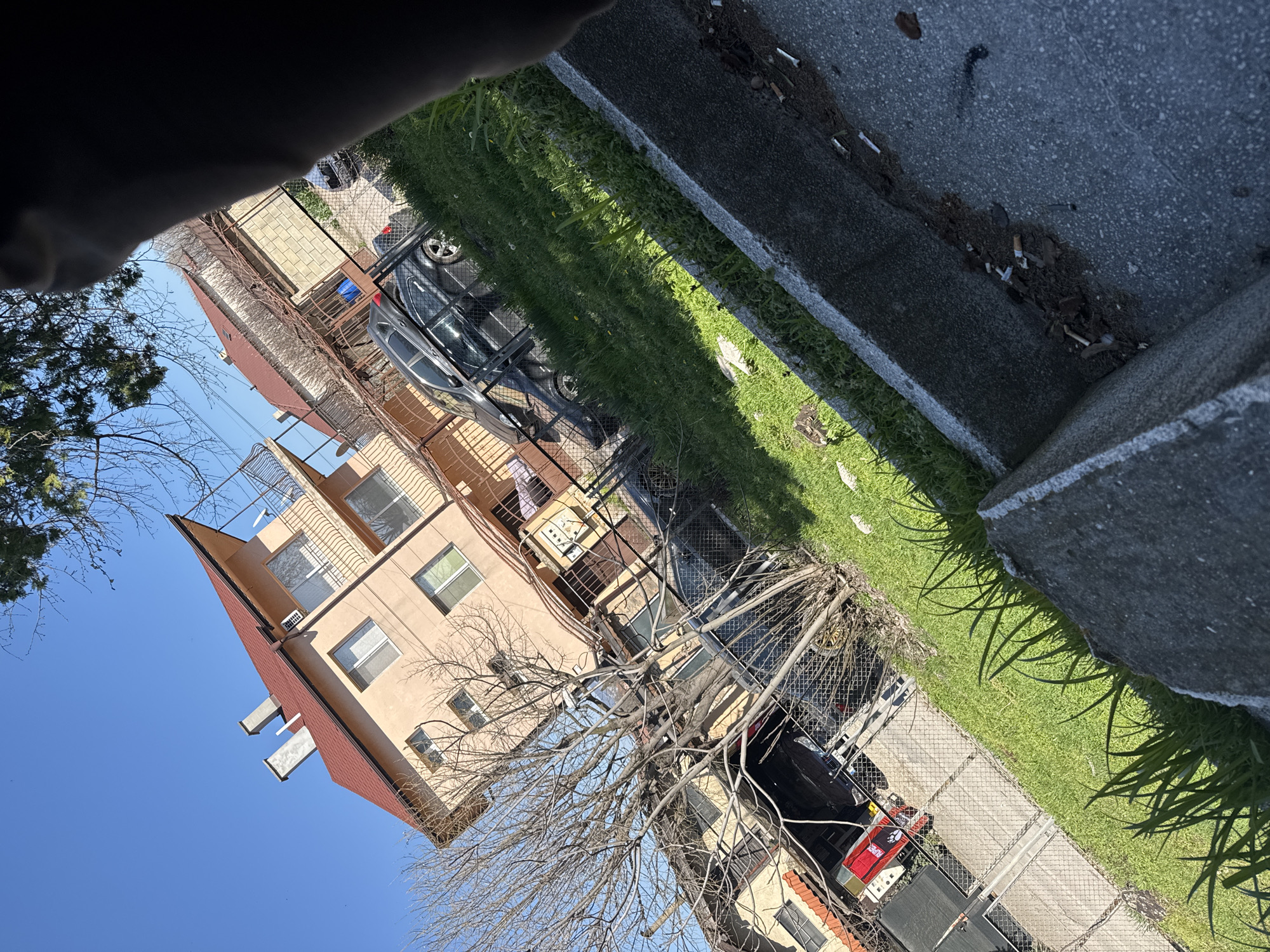 The image shows a residential scene, possibly in Topoli, Bulgaria, featuring a light peach-colored building with a red roof. The building is positioned next to a chain-link fence. A dark-colored car is parked behind the fence, with a tree's branches obscuring part of the view. Patches of green grass are visible between the fence and a concrete curb. Discarded cigarette butts are scattered along the edge of the curb. The sky is blue, and a partial view of a person's head is visible in the upper left corner of the frame.