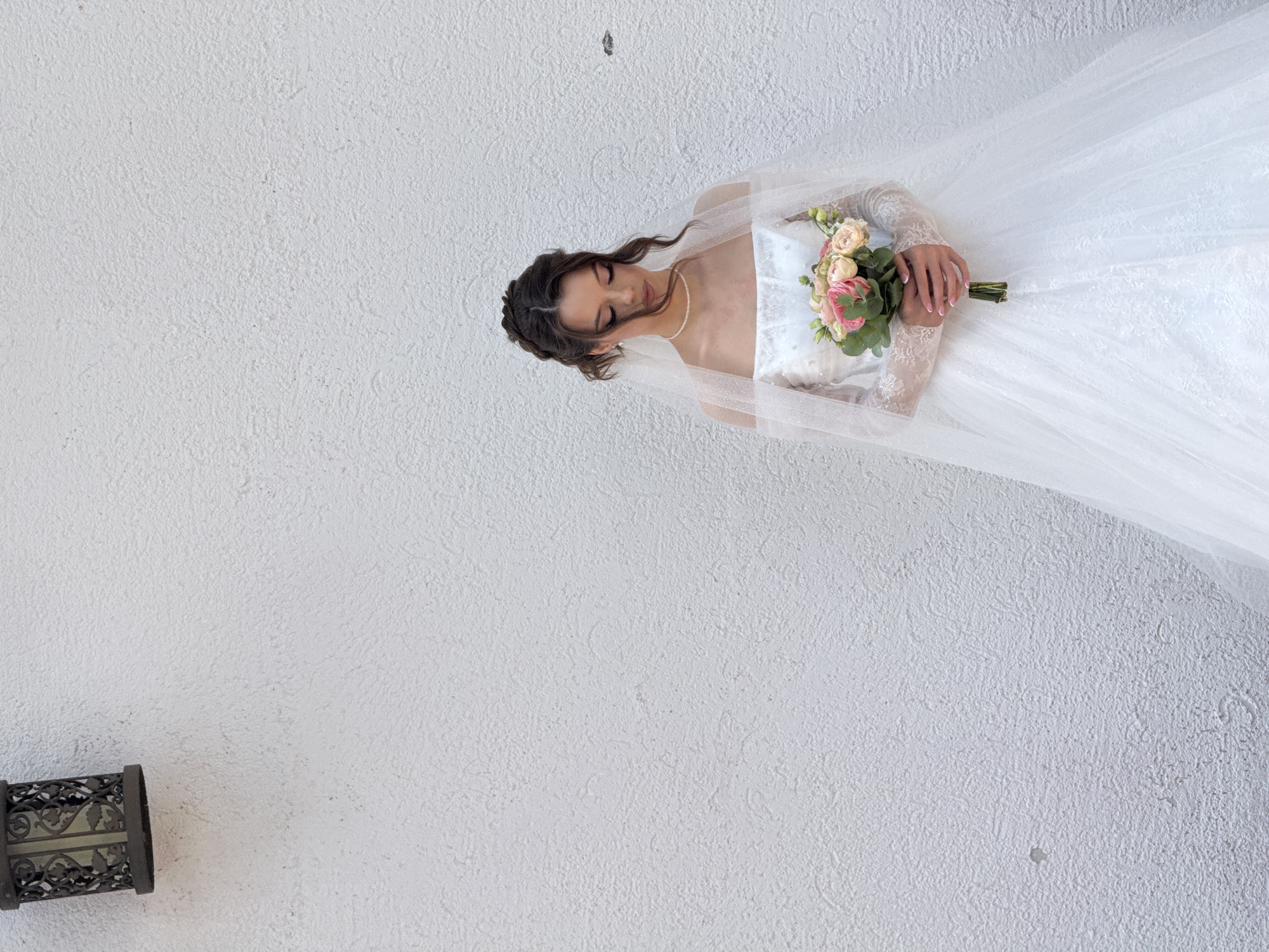 A young woman, dressed in a white wedding gown and veil, stands against a textured white wall, holding a bouquet of pink and yellow roses. Her eyes are downcast, and her hair is styled in an updo. A decorative lantern is mounted on the wall to the upper left of the frame. The scene appears to be outdoors, possibly in a courtyard or garden. The image's composition suggests a wedding portrait. The location is not discernable from the image itself, and there is no visual information to indicate it is in Votkinsk, Russia.