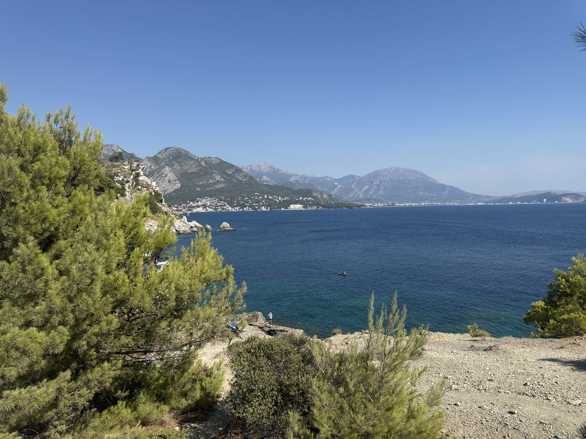 The image shows a scenic view of the Adriatic Sea and a coastline with mountains in the distance. In the foreground, there are pine trees and rocky terrain. The sea is a deep blue, and the sky is clear. On the coastline, there are buildings and settlements nestled among the green hills. A small boat or kayak is visible in the water. Closer to the shore, a person is standing on rocks, possibly fishing or observing the sea. There is also a second person in the water, possibly swimming or snorkeling. The overall scene depicts a tranquil coastal landscape, likely in the vicinity of Sutomore, Montenegro.