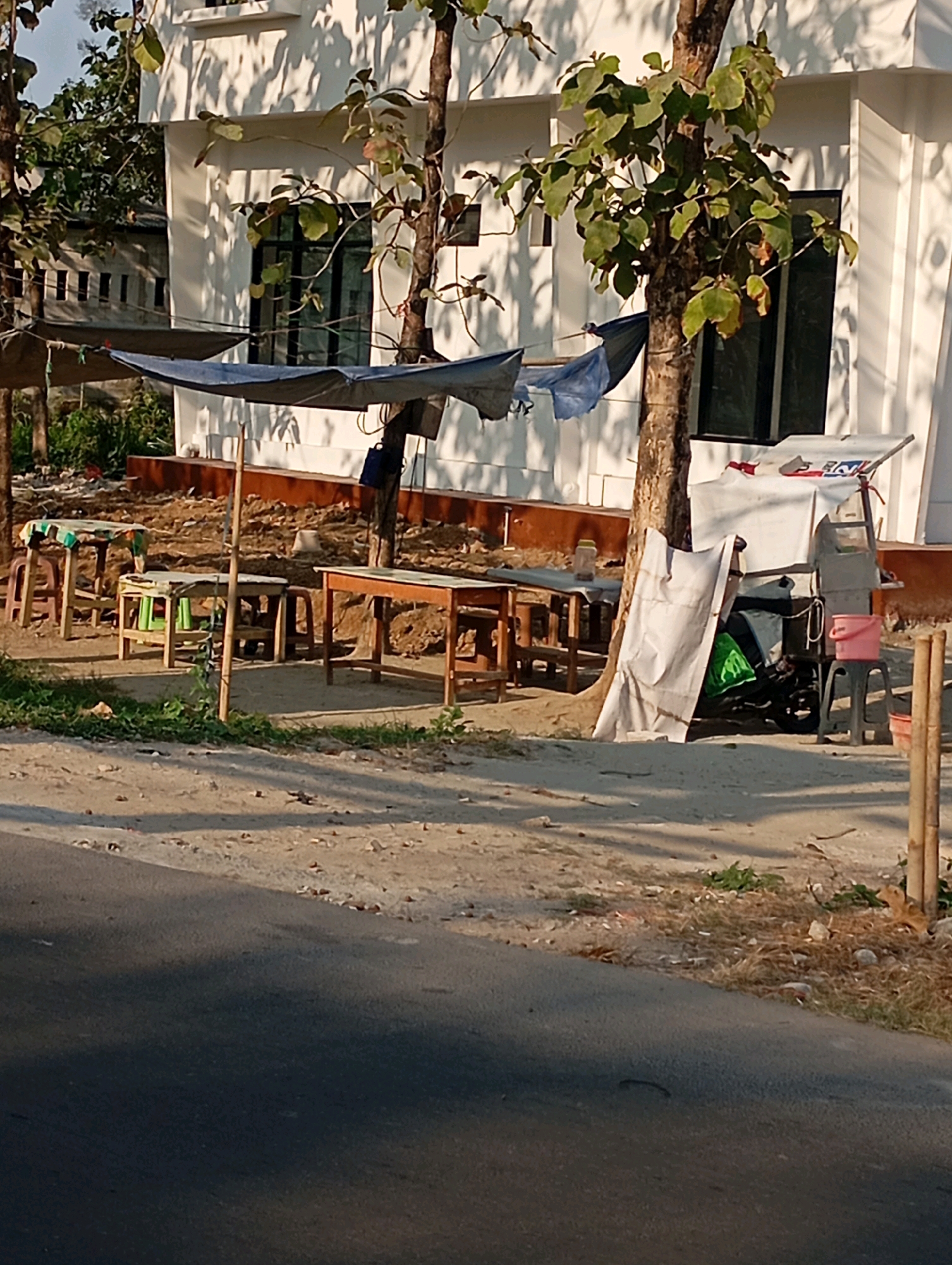 The image depicts an outdoor scene in Babelan, Indonesia, featuring a white building with dark-framed windows. In front of the building, a temporary outdoor seating area is set up with several wooden tables and chairs. Some of these tables are covered with cloth. A blue tarp is stretched between trees, providing shade over a portion of the seating area. To the right, there is a small cart or stand covered with white fabric, and next to it, a pink bucket sits on a black stool. A green object, possibly a bag or cushion, is visible under the white covering. The ground is a mixture of sand and dry vegetation, with scattered debris. A paved road is visible in the foreground, with shadows cast across it and the surrounding ground, indicating daylight. The overall impression is of a simple, open-air food or rest stop.