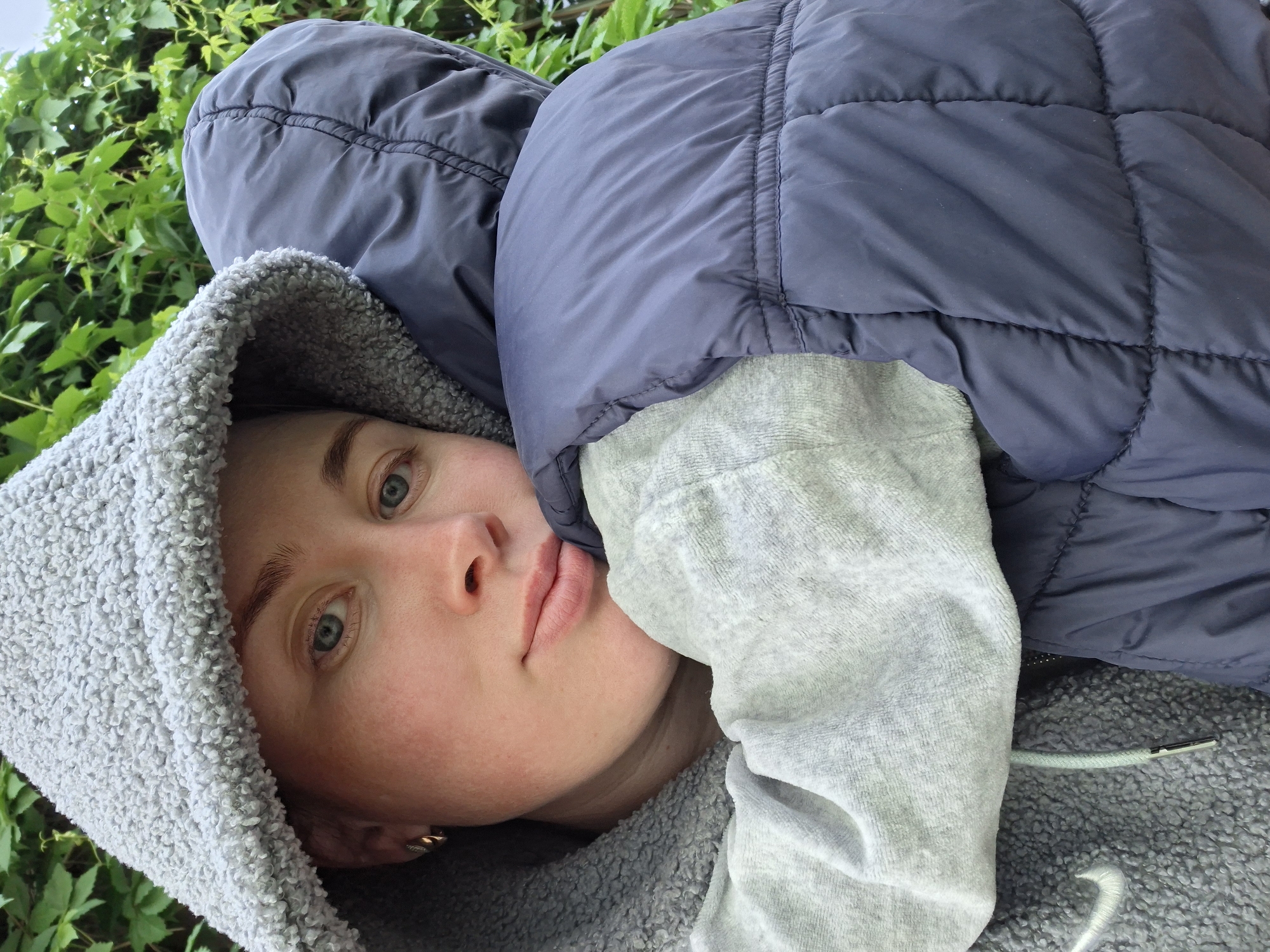 A young adult woman with light skin and blue eyes is viewed from a low angle, lying down against a background of green foliage. She is wearing a grey hooded sweatshirt, and the hood is up. A puffy, navy blue jacket covers most of her body. The location is in the city of Unknown, Unknown.