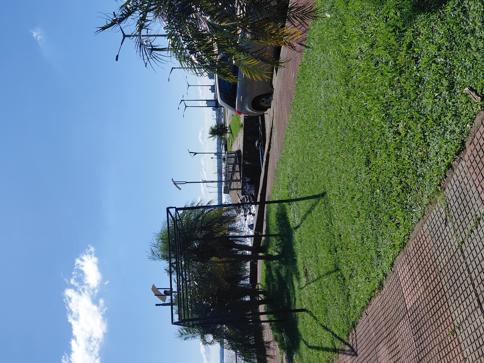 The image depicts a waterfront park area in Encarnación, Paraguay, on a sunny day with a clear blue sky and scattered white clouds. In the foreground, a paved walkway made of reddish-brown square tiles runs along the edge of a well-maintained green lawn. A skeletal metal structure, possibly a canopy or trellis, stands on the lawn, casting shadows. Several palm trees with distinct fronds are clustered behind the structure, their leaves rustling slightly.
Beyond the lawn and palm trees, a body of water, likely the Paraná River, stretches across the background, its surface shimmering under the sunlight. A concrete embankment and walkway run parallel to the water. Several lampposts line this walkway. A silver SUV is parked on the embankment near the water's edge, partially obscuring a trailer. In the distance, more lampposts and faint city structures can be seen along the waterfront, suggesting an urban setting. The overall impression is one of a public recreational space by the water, enjoying pleasant weather.