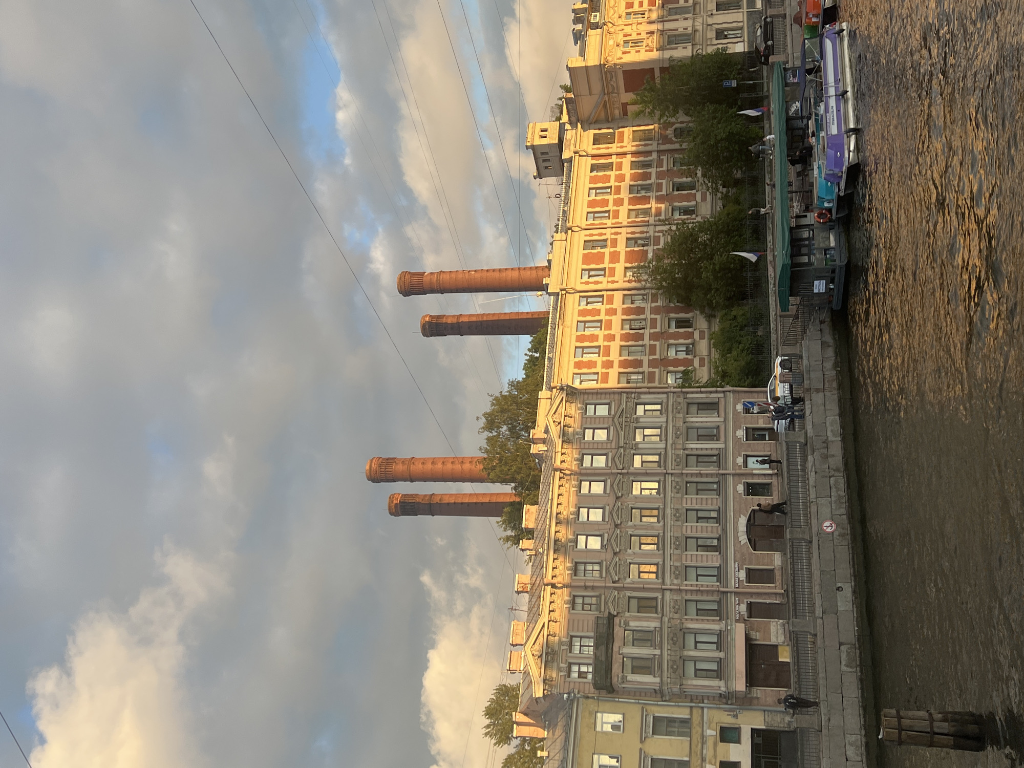 The image shows a view of a riverfront in Saint Petersburg, Russia, likely during the late afternoon or early evening due to the warm, golden light reflecting off the buildings and water. The scene features several industrial-style brick buildings with numerous windows, characteristic of historic urban architecture in the city. Prominently in the foreground are three large, rusty brick smokestacks, suggesting a former industrial area.
Along the riverbank, there are several boats docked. One is a modern pleasure boat with a purple and white design, others are less distinct but appear to be covered for protection. A few people are visible on the embankment, some walking, one near a boat, and others further in the distance, their presence adding a sense of scale and activity to the urban landscape. Trees line parts of the embankment, providing a touch of greenery against the brick and water.
Overhead, thick clouds are present, tinged with the warm light, indicating a partly cloudy sky. Electrical wires are strung across the upper portion of the frame, a common feature in urban environments. The water of the river is choppy, with ripples catching the light, and shows a murky, brownish hue.
There is some visible text on a sign on one of the boats, but it is illegible. The overall mood of the image is one of historical industrial grandeur meeting modern urban life, bathed in the soft light of a late day.