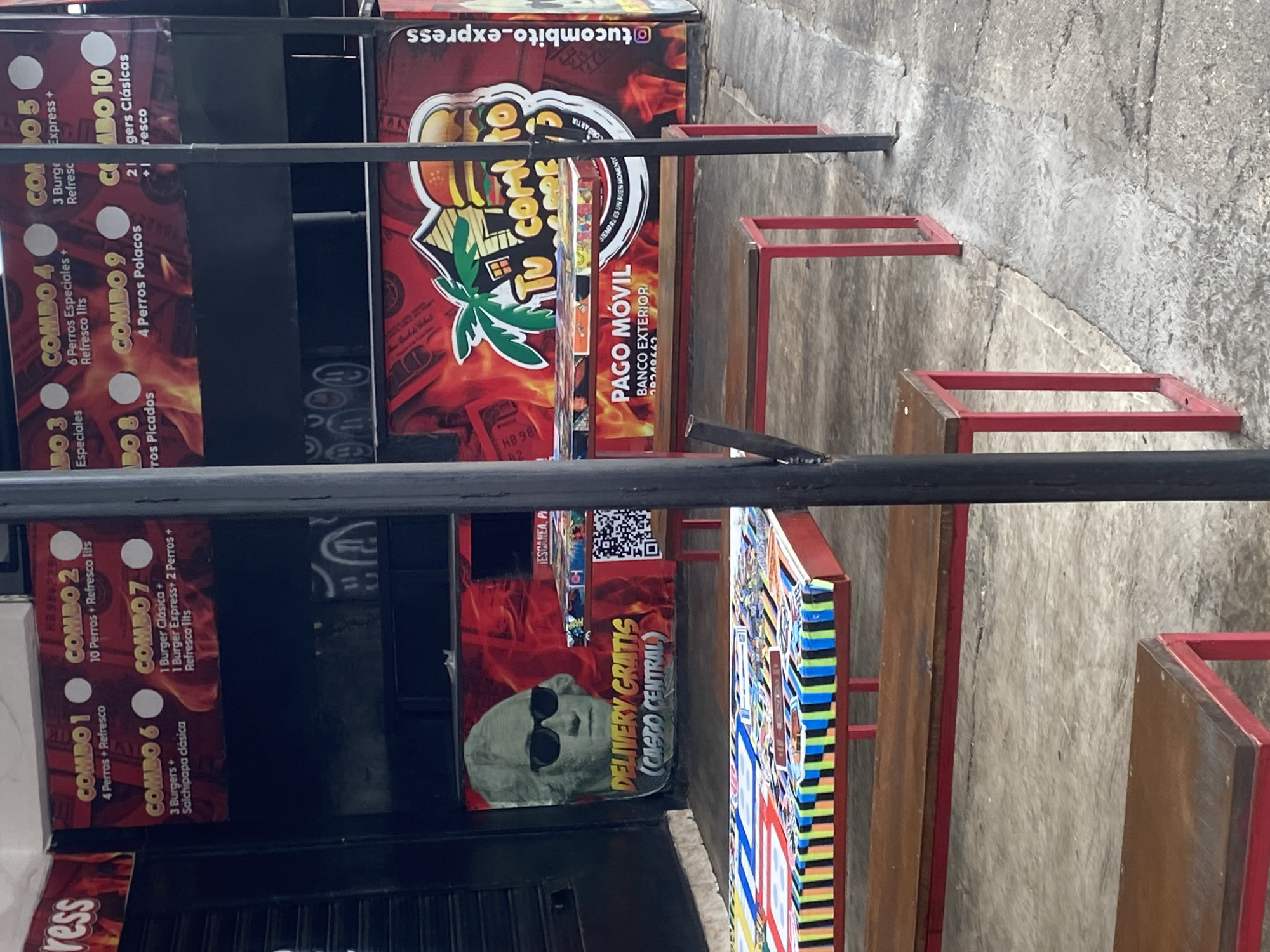 A low-angle shot depicts a street food stall in Tinaquillo, Venezuela. The stall features advertising boards with the menu offering "Combos" of hot dogs and burgers. One board advertises "Delivery Gratis (Casco Central)" alongside an image of George Washington wearing sunglasses. The menu is written in Spanish. Behind the stall, a narrow walkway is visible, constructed of concrete. Several small tables with red frames and wooden surfaces are visible, suggesting seating for customers.