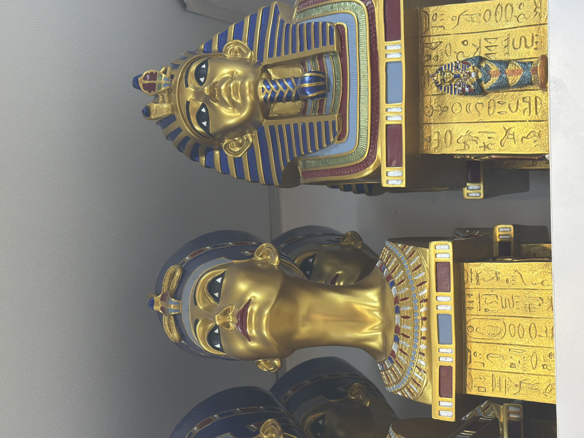 This is a close-up, eye-level, indoor shot of two Egyptian-themed busts displayed on a shelf. The bust on the left is of a female figure, likely Nefertiti, with a golden face, wearing a blue and gold headdress. She has her head tilted upwards and is looking to the left. The bust on the right is of a male figure, likely a pharaoh, also golden, with a blue and gold striped headdress. This bust is positioned on a larger, rectangular base with hieroglyphics and a smaller sarcophagus-like figure on its front. The background is a plain, light-colored ceiling or shelf. The overall impression is that these are decorative items, possibly souvenirs, given the context of Sharm El Sheikh, Egypt, which is a popular tourist destination known for its Egyptian heritage and souvenir shops.