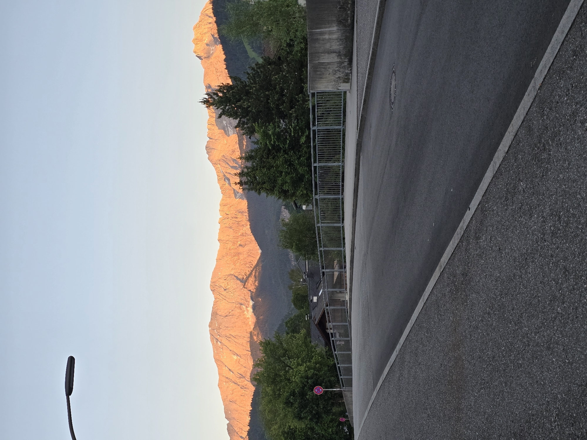 The image depicts a view of the Zugspitze mountain range illuminated by the golden light of sunrise, seen from a street in Garmisch-Partenkirchen, Germany. In the foreground, a paved road runs diagonally from the bottom left to the top right of the frame. The road is bordered by a light-colored curb and a darker asphalt surface. On the left side of the road, a light gray sidewalk with a textured surface is visible. A street lamp with a dark, elongated fixture extends from the upper left corner, casting no illumination in the bright morning light. Beyond the road, a metal railing runs along the edge of a raised area. A concrete wall is situated to the right of the railing. In the middle ground, a line of coniferous trees stands, interspersed with deciduous trees and some buildings with dark roofs. A red and white circular sign with two arrows is visible on the left side, near the railing. The background is dominated by the majestic Zugspitze mountain range, its peaks glowing with the warm hues of sunrise, with patches of snow visible on the upper slopes. The sky is a clear, pale blue. The overall impression is one of a tranquil morning scene in a mountainous region.