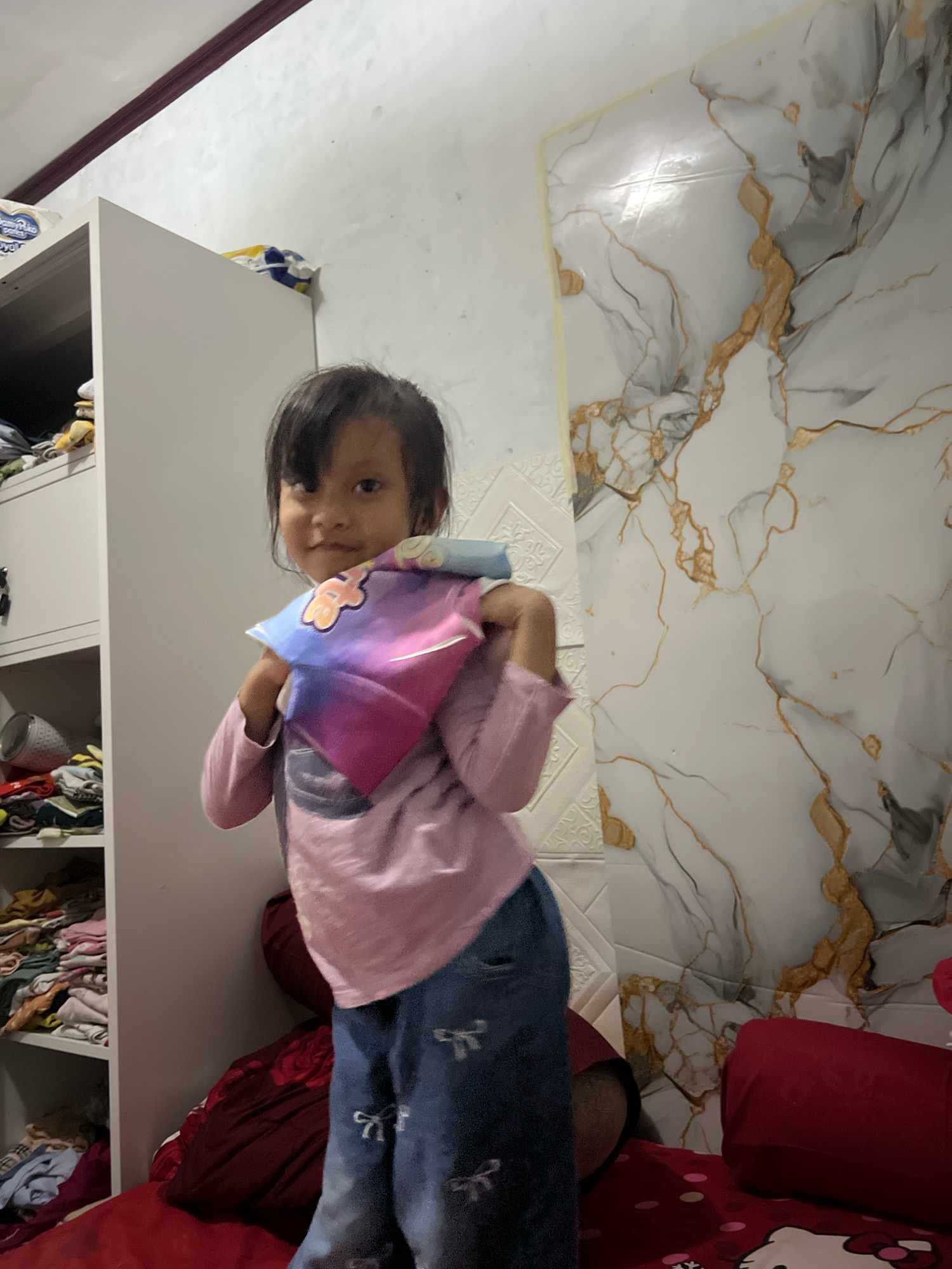 A young girl, appearing to be of East Asian descent, stands in a room. She has dark hair, a light complexion, and is wearing a pink long-sleeved shirt and blue jeans with white bow patterns. She is holding a colorful, folded fabric, possibly a scarf or a toy, in front of her chest with both hands.
The background features a white wardrobe on the left, filled with neatly folded clothing on its shelves. To the right of the wardrobe and behind the girl, there is a wall with a decorative marble-like pattern in white, grey, and gold. The floor appears to be covered with red bedding, and a red bolster pillow is visible.
The lighting in the room is bright, suggesting an indoor setting during the daytime. The overall impression is of a domestic interior, possibly a bedroom or a play area.
The provided location context places this scene in Kecamatan Pati, Indonesia, indicating the specific geographical area where the photograph was taken.