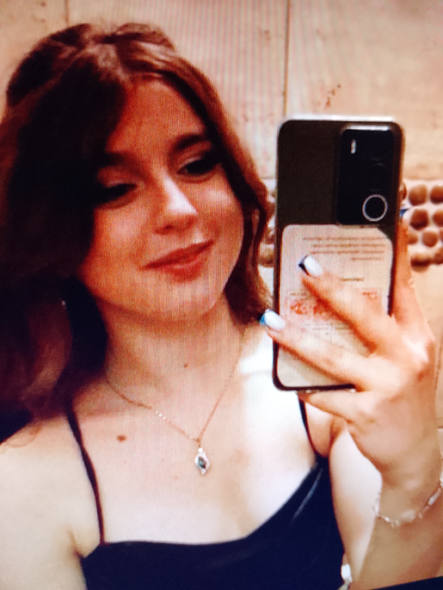 A young woman with long, dark brown hair, wearing a black spaghetti strap top and a delicate necklace with a light-colored pendant, is taking a selfie in an indoor setting. She is holding a smartphone in her right hand, which is positioned to capture her reflection. Her nails are painted white with blue tips. The background appears to be a wall with a textured, tiled surface. The image quality is low, with visible scan lines, suggesting it may be a photograph of a screen. The location is identified as Ostróda, Poland.