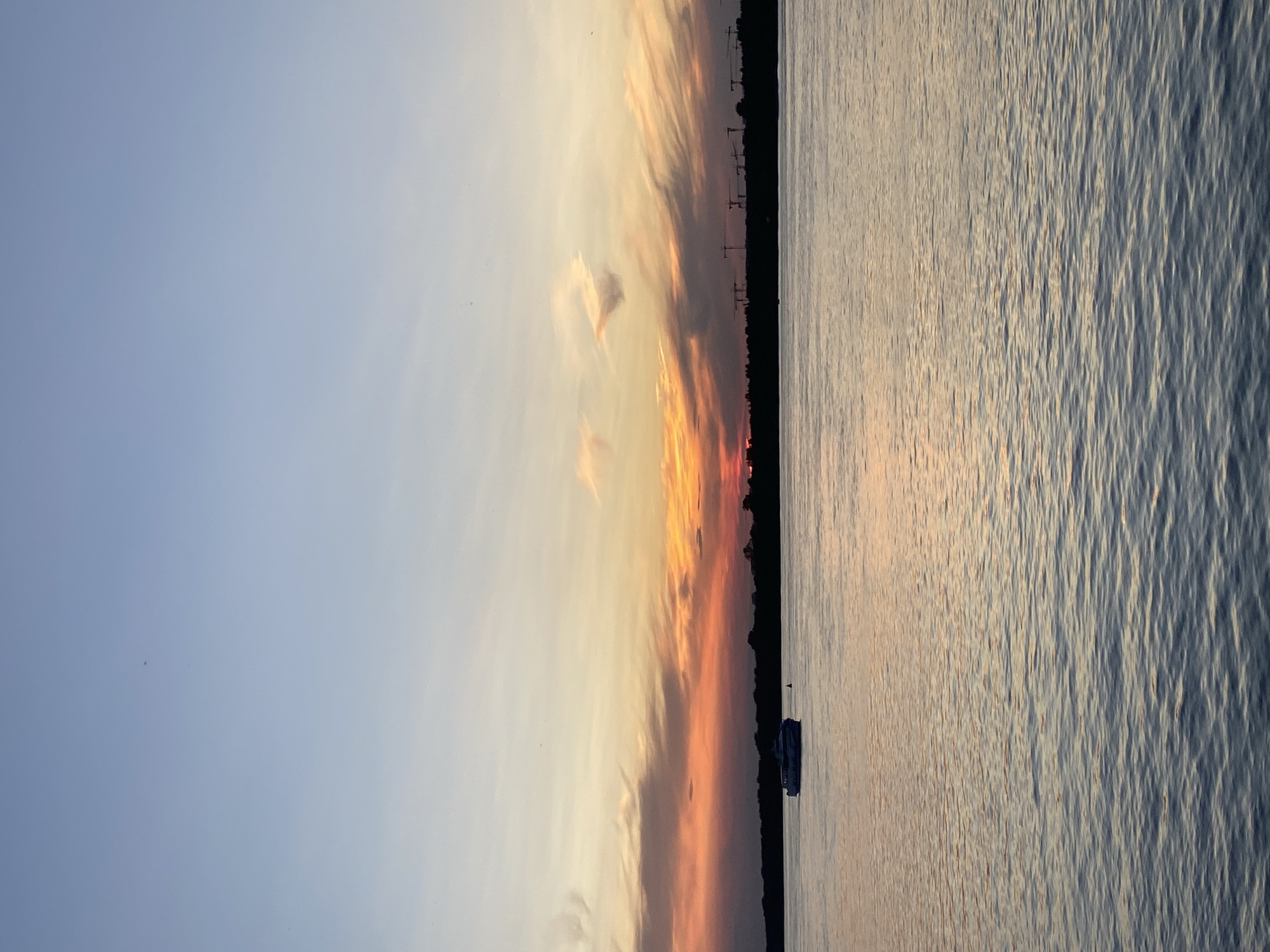 The image captures a sunset over a body of water, likely the Kama River in Perm, Russia. The sky is a gradient of blue, transitioning to hues of orange and red near the horizon, with wispy clouds. A silhouette of trees and construction cranes is visible along the distant shoreline. In the foreground, a small boat sails on the calm water, reflecting the colors of the sunset. The overall scene is serene and tranquil.