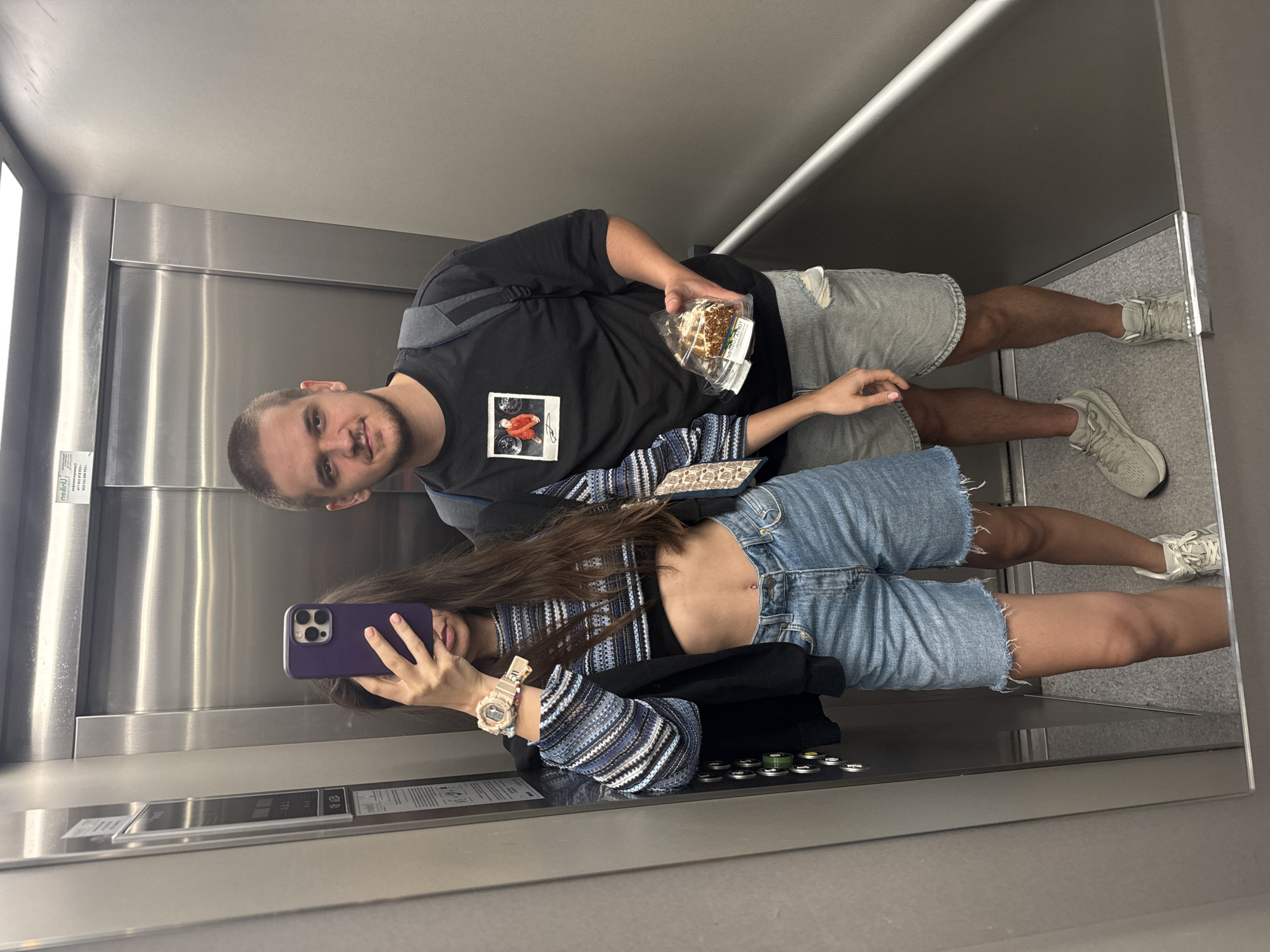 Two individuals are visible in an elevator. A man wearing a black t-shirt with an image on it, gray shorts, and sneakers is holding a clear container with food. A woman is holding a purple cell phone, taking a selfie. She is wearing denim shorts, a black top with a striped jacket, and a watch. The interior of the elevator has metal walls and controls. This image appears to be taken in an elevator in the city of Plovdiv, Bulgaria.