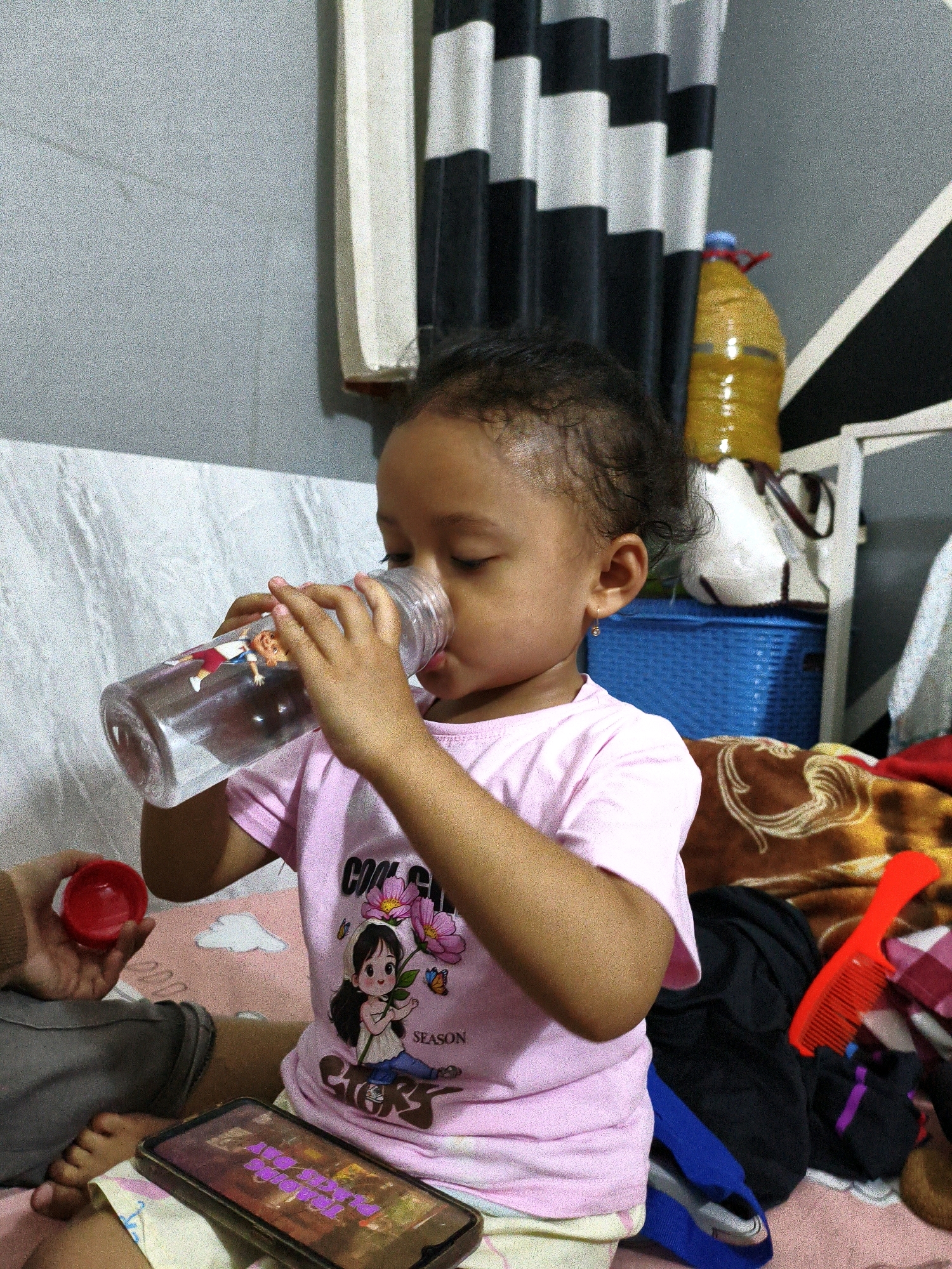A young child, likely female based on clothing and accessories, is seated on a pink surface, possibly a bed or mat, in a room. The child is drinking from a clear plastic water bottle with cartoon decals. She wears a pink t-shirt with a graphic design of a girl holding flowers. In her left hand, she holds a small red object, possibly a bottle cap. To the left of the child, a portion of a person's leg, covered in gray fabric, is visible. The background reveals a black and white striped curtain, a yellow bottle with a red cap, and a blue woven basket. A brown and tan patterned blanket is draped behind the child, and a red comb lies on top of some dark fabric. A smartphone with a screen displaying colorful text is placed in front of the child. The overall impression is a candid, indoor scene, likely in a residential setting. The image is grainy, suggesting it was taken with a low-light or older camera.