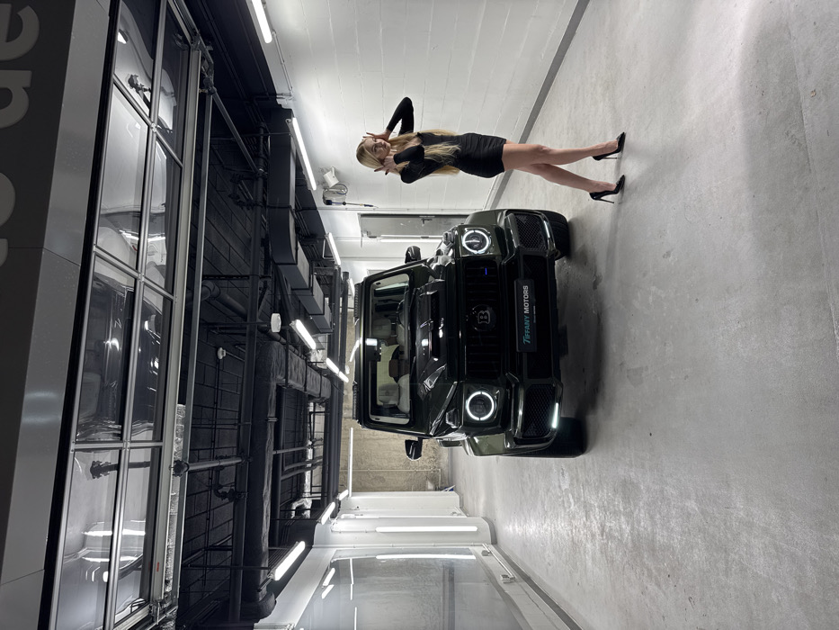 The scene depicts the interior of what appears to be a luxury automotive facility, identified as Tiffany Motors in Warsaw, Poland, based on the license plate frame. A dark green, customized Brabus Mercedes-Benz G-Wagen is parked centrally, facing the camera, with its distinctive circular LED daytime running lights illuminated. To the vehicle's right, a blonde female wearing a black long-sleeved mini-dress and black high heels stands posing, with her hands positioned near her head. The environment features a smooth, light gray concrete floor, white walls, and an industrial-style ceiling with exposed black-painted ducts, pipes, and fluorescent lighting fixtures. Vertical LED light panels are visible on the left wall, and a garage door with translucent panels is partially open overhead. A white door is present on the right wall.