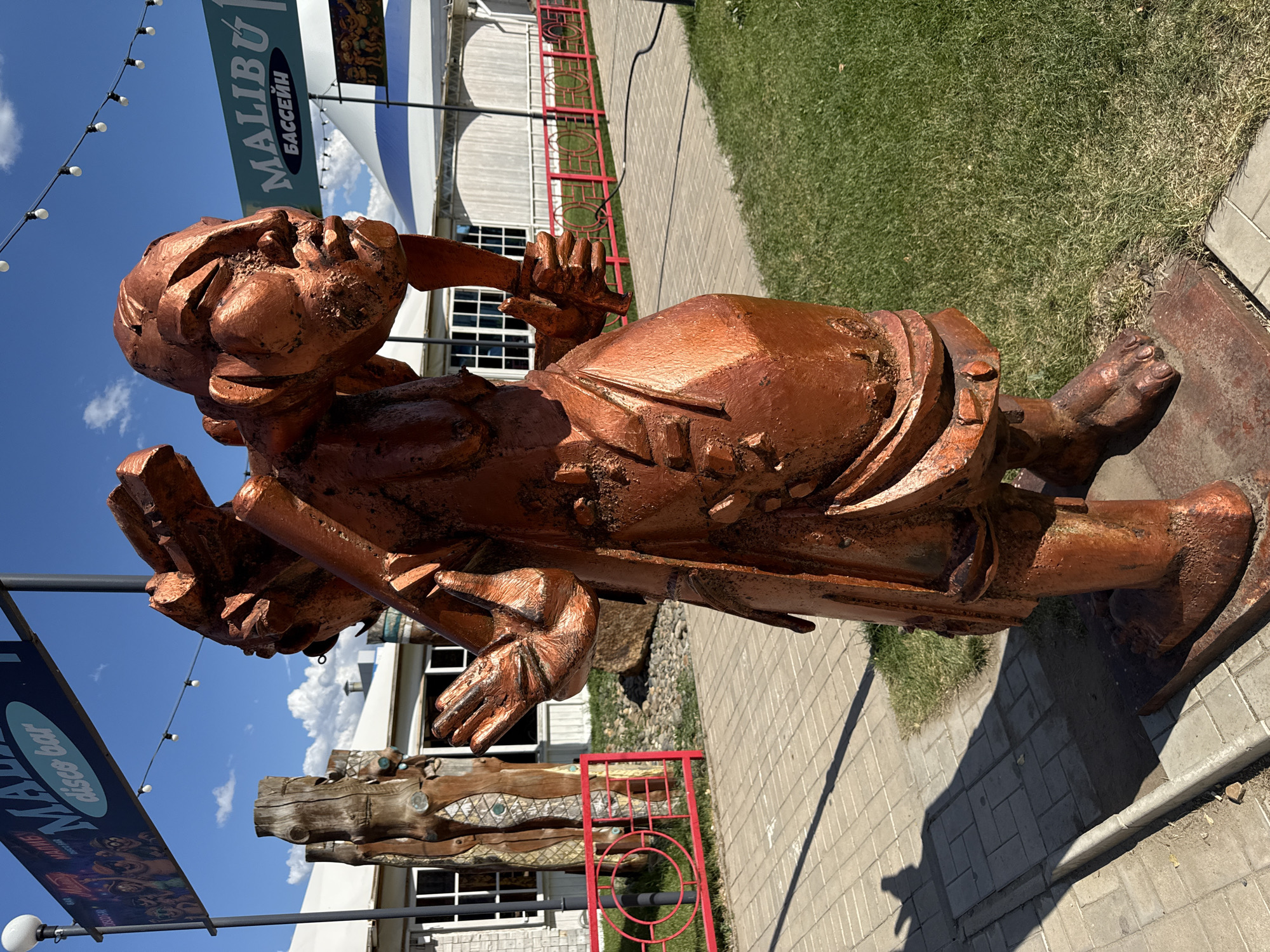 A full shot captures a weathered, copper-colored statue of a person in an outdoor setting, likely in Yarovoye, Russia. The statue depicts a figure from the waist up, with a distinctly Asian-inspired facial structure, large hands, and a robe-like garment. The figure is carrying a large load on its back and holds a tool or weapon in its right hand, while its left hand is raised in a gesture of greeting or warning. The statue is positioned on a paved walkway, with a strip of green grass to its right. In the background, a building with large windows and a white roof is visible, along with a red metal fence. Overhead, a clear blue sky is dotted with fluffy white clouds, and a string of lights hangs across the scene, suggesting a recreational or entertainment area. A banner for "Malibu Disco Bar" is also visible in the background. The lighting indicates it is daytime.