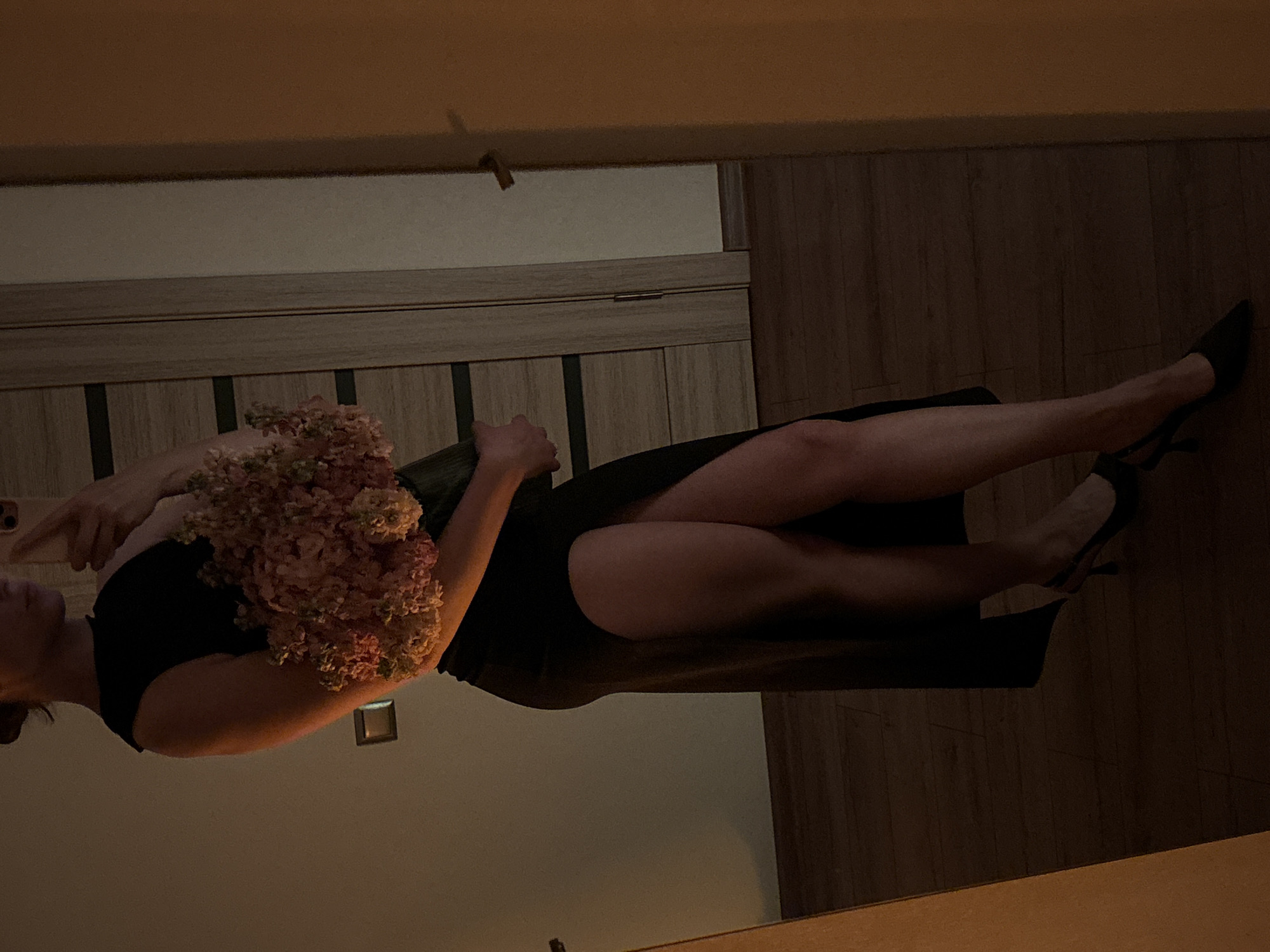 A person, seen from the mid-torso down, stands in what appears to be an indoor setting, possibly a room in a home. They are holding a bouquet of pink and white flowers. The person is wearing a black dress with a high slit on the left side, revealing their legs. They are also wearing black slingback heels. The lighting in the room is dim, with a warm glow, and the scene is reflected in a mirror. The background shows a wooden floor and a door with horizontal paneling. The overall impression is one of a posed photograph taken in low light conditions.