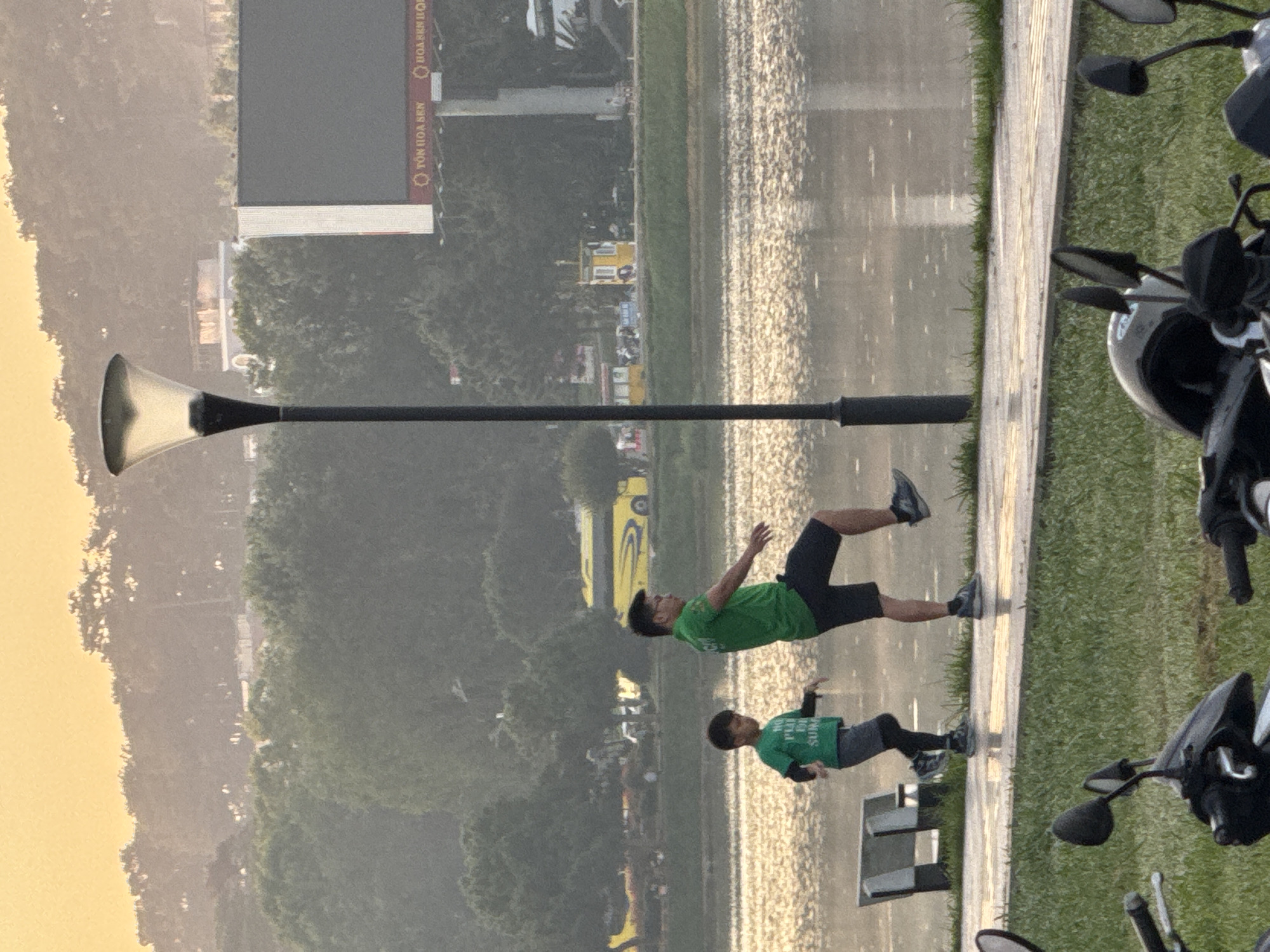 A father and son are practicing tai chi by Xuan Huong Lake in Da Lat, Vietnam. The father, wearing a green t-shirt and black shorts, is in mid-stride with his right leg extended forward. His son, in a green t-shirt and grey shorts, mimics his movements with his left leg forward. The lake's surface reflects the golden light of the setting sun, creating a shimmering effect. In the background, trees line the shore, and a yellow bus is visible on a road. A tall lamppost stands prominently on the right, and a billboard with Vietnamese text is also present. Motorcycles are parked in the foreground.