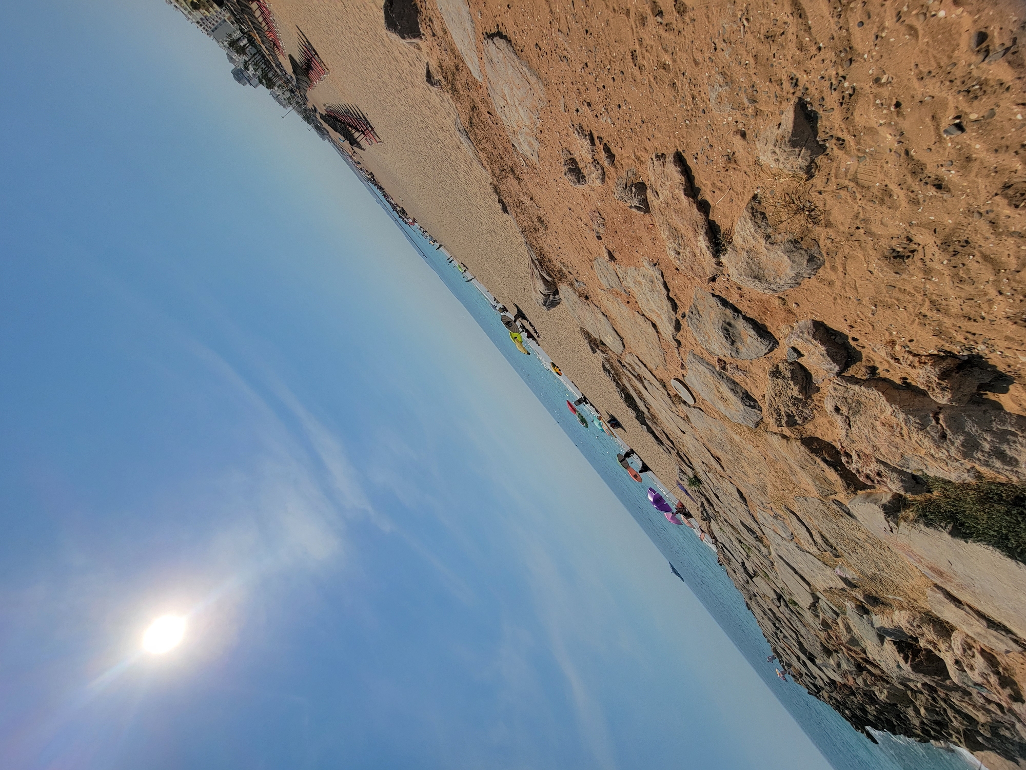 The image depicts a sunny day at a beach in Quarteira, Portugal. The scene is dominated by a wide expanse of sandy beach and the calm, blue sea. In the foreground, a rocky embankment lines the edge of the beach. Scattered across the sand are numerous beach chairs and umbrellas, indicating a popular spot for sunbathers.
Several people are visible along the shoreline, some walking, and others appearing to be engaged in activities near the water. A few colorful umbrellas are set up on the beach. The sun is bright and prominent in the clear blue sky, suggesting it is midday. The overall mood is relaxed and leisurely, characteristic of a beach vacation.
The composition of the image is striking, with the beach and sea stretching out diagonally across the frame, from the upper left to the lower right. The rocky texture of the embankment in the foreground provides a contrast to the smooth sand and water. The clear sky and bright sunlight indicate fair weather. No visible text is present in the image.