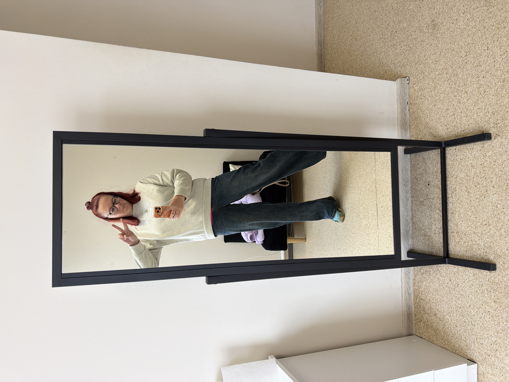 A mirror reflects a woman with reddish hair and glasses, holding a phone in her right hand and making a peace sign with her left. She wears a light-colored sweater and dark denim jeans, with a bit of a pink or red garment visible under her sweater. She stands in what appears to be a room with light beige textured flooring and white walls. The mirror is rectangular, framed in black metal, with a stand that has thin metal supports. Behind the woman, visible in the mirror's reflection, there is a black chair and what looks like some clothes or fabric draped on it. It is not possible to confirm if the image was taken in Chaikovsky, Russia.