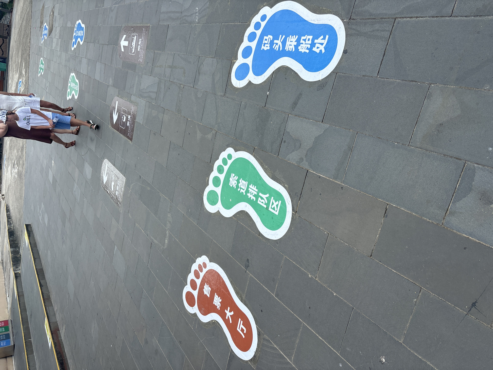 The image depicts a paved walkway in Xincun, China, marked with colorful footprints indicating directional queues. Three prominent footprints are visible in the foreground: a red one on the left with Chinese characters "售票大厅" (Ticket Hall), a green one in the center with "索道排队区" (Cable Car Queue Area), and a blue one on the right with "码头乘船处" (Pier Boarding Area). In the background, other smaller footprints and directional signs for the Cable Car Station and Tourist Ferry Terminal are also present. A family, including a young boy wearing a white t-shirt that says "CHILL," is walking away from the camera, passing by some of the directional markers. The walkway is paved with dark gray square tiles, and there is a raised concrete border with yellow markings along the left side of the image. The environment appears to be outdoors, possibly at a tourist attraction or transportation hub.