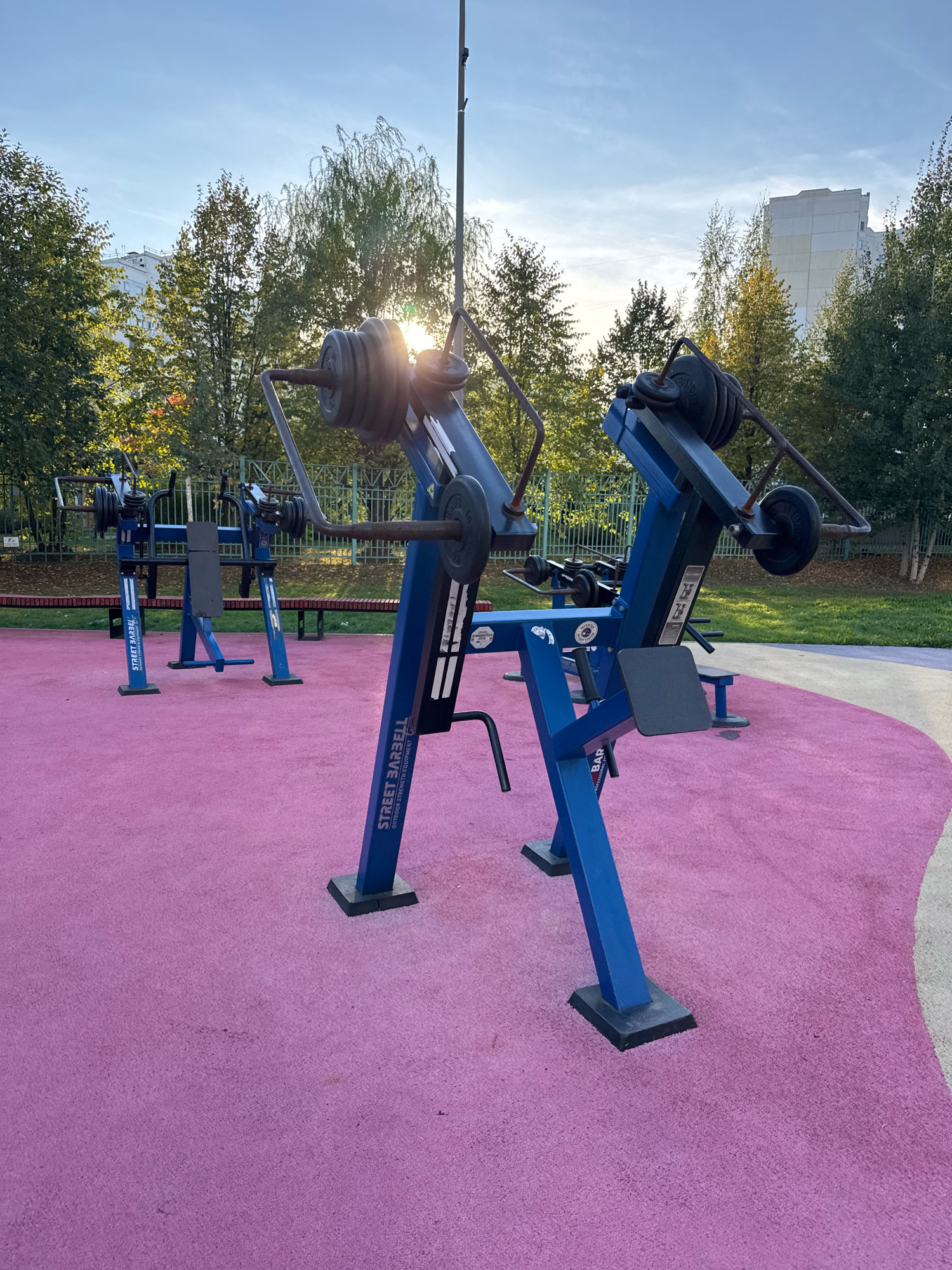 The image depicts an outdoor gym area with several pieces of blue metal exercise equipment. The equipment appears to be weight machines with visible weight plates. The ground surface is a textured pink material. In the background, there are trees with green and yellowing leaves, suggesting it is autumn. Behind the trees, a tall, light-colored building is visible, as well as a clear sky with the sun shining through the trees, creating lens flare. A metal fence is also present in the background. The scene is located in Милицейский посёлок, Russia. There are no people visible in the image.
