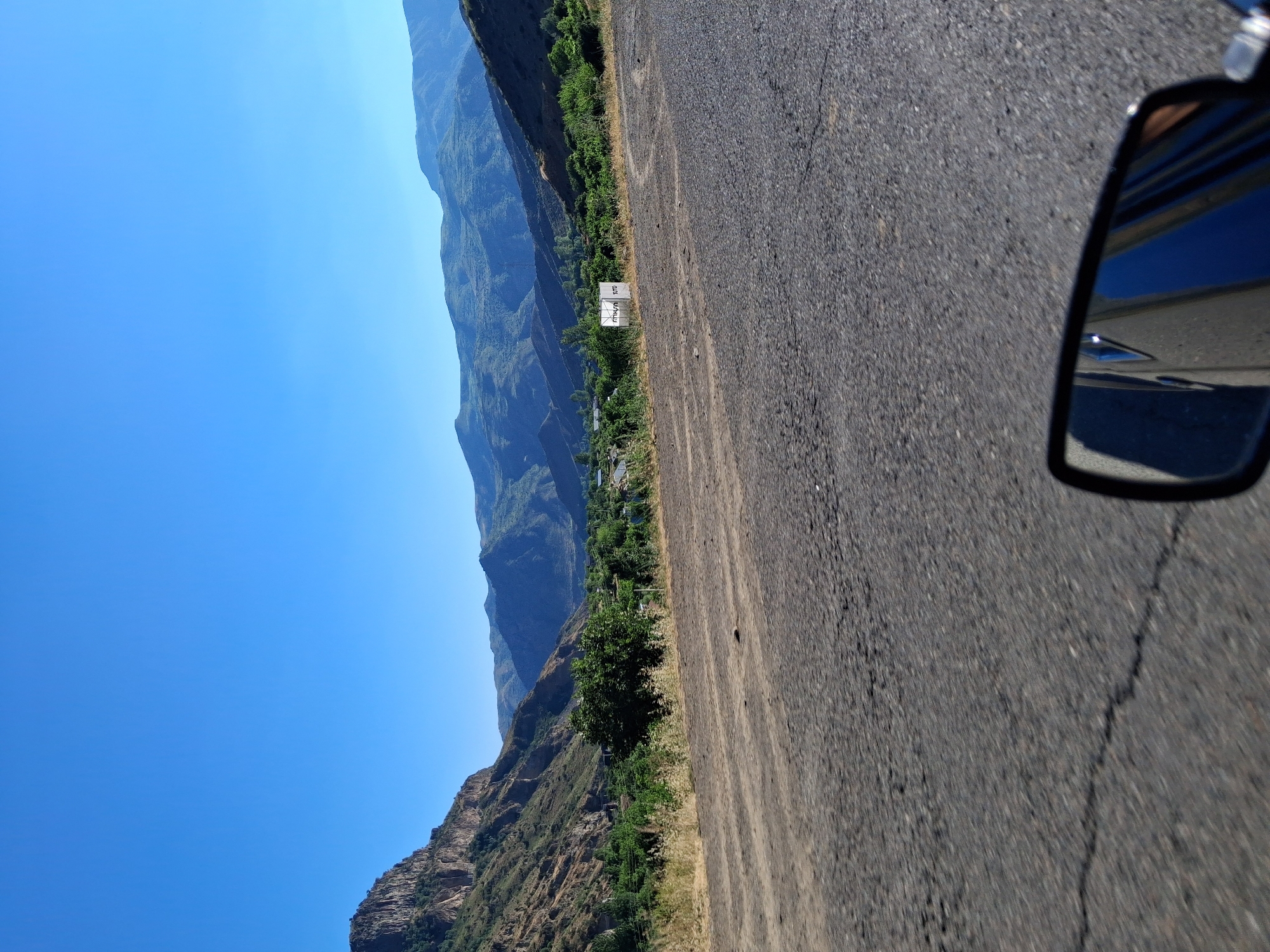 This media file captures a scenic view of a mountainous landscape with a clear blue sky. The perspective suggests the image was taken from a vehicle, as a car's side mirror is visible in the lower right corner. The road itself appears to be asphalt with some visible cracks and patches.
To the left of the road, a steep, rugged mountain range dominates the scene, covered in patches of green vegetation. The mountains stretch into the distance, creating a sense of depth and vastness. Below the mountains, a small settlement or village is visible, nestled amongst trees and greenery. A white sign with some text is also present near the roadside, though the text is not clearly legible.
The lighting and the bright blue sky suggest it is daytime, likely morning or afternoon. The weather appears to be clear and sunny, ideal for driving through this mountainous region. The location cues, such as the style of the mountains and the vegetation, are consistent with a setting in Shatin, Armenia. There are no people or specific activities depicted, emphasizing the natural beauty of the landscape. The overall mood is one of tranquility and exploration.