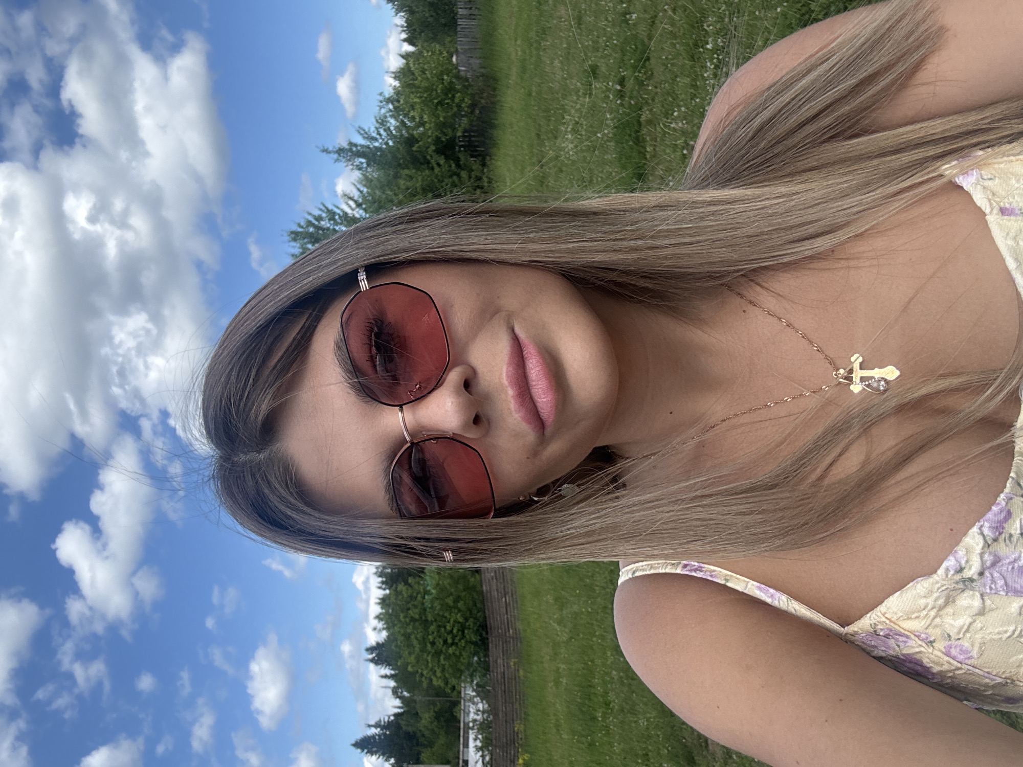 A young woman with long, blonde hair and pink lipstick smiles as she takes a selfie outdoors. She wears pink-tinted sunglasses and a floral-patterned dress. A delicate gold necklace with a cross pendant rests on her collarbone. The background shows a lush green field, a treeline, and a bright blue sky with scattered white clouds, suggesting a sunny day. A rustic wooden fence is visible in the distance. The overall mood is relaxed and cheerful, indicative of a pleasant outdoor leisure activity. The image appears to have been taken during daylight hours, possibly in the afternoon, given the bright sunlight and clear sky. The location context is Пастухово, Russia. There is no visible text in the image.